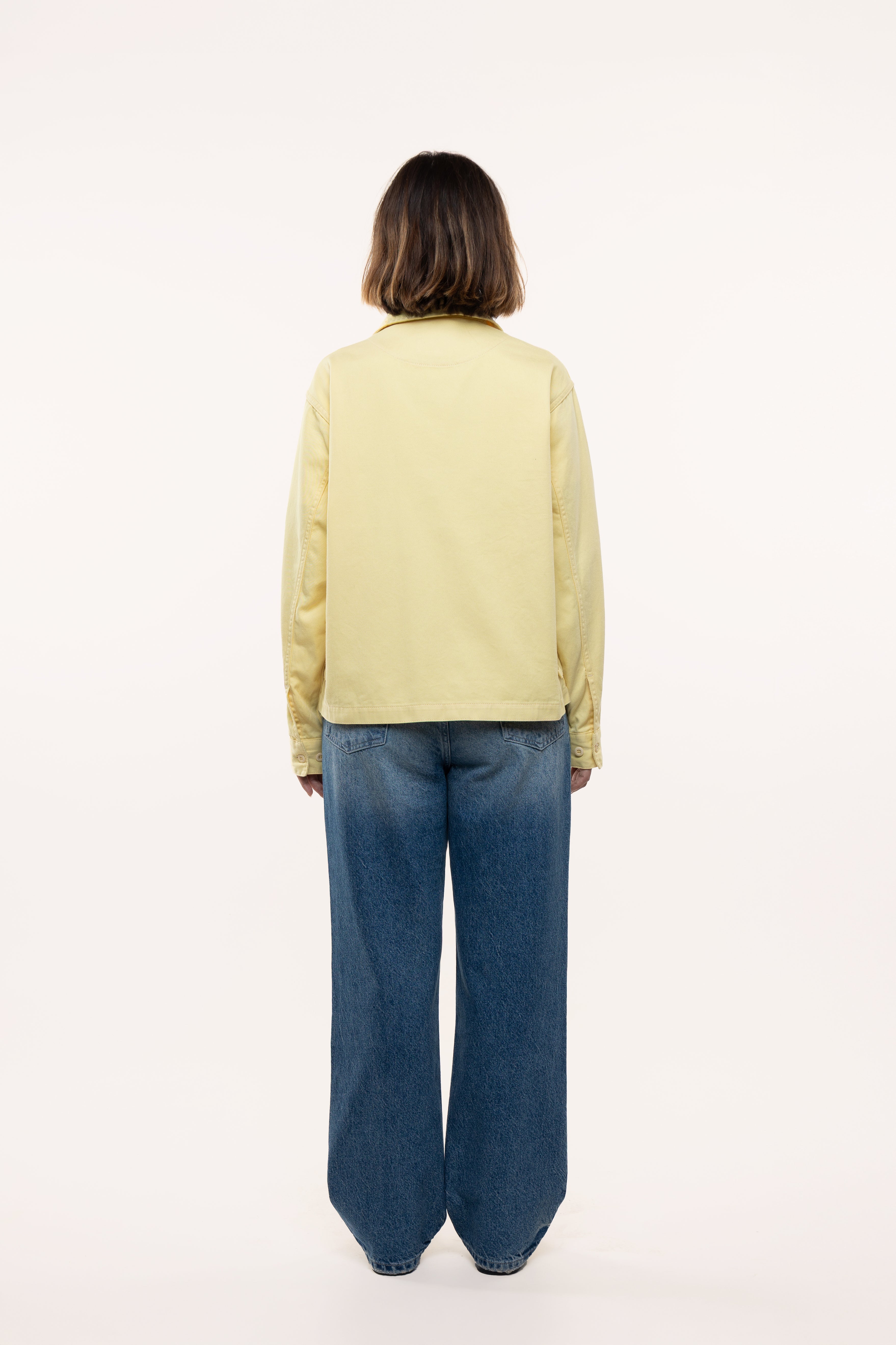 Back view of a woman wearing a light green jacket and blue jeans on a white background