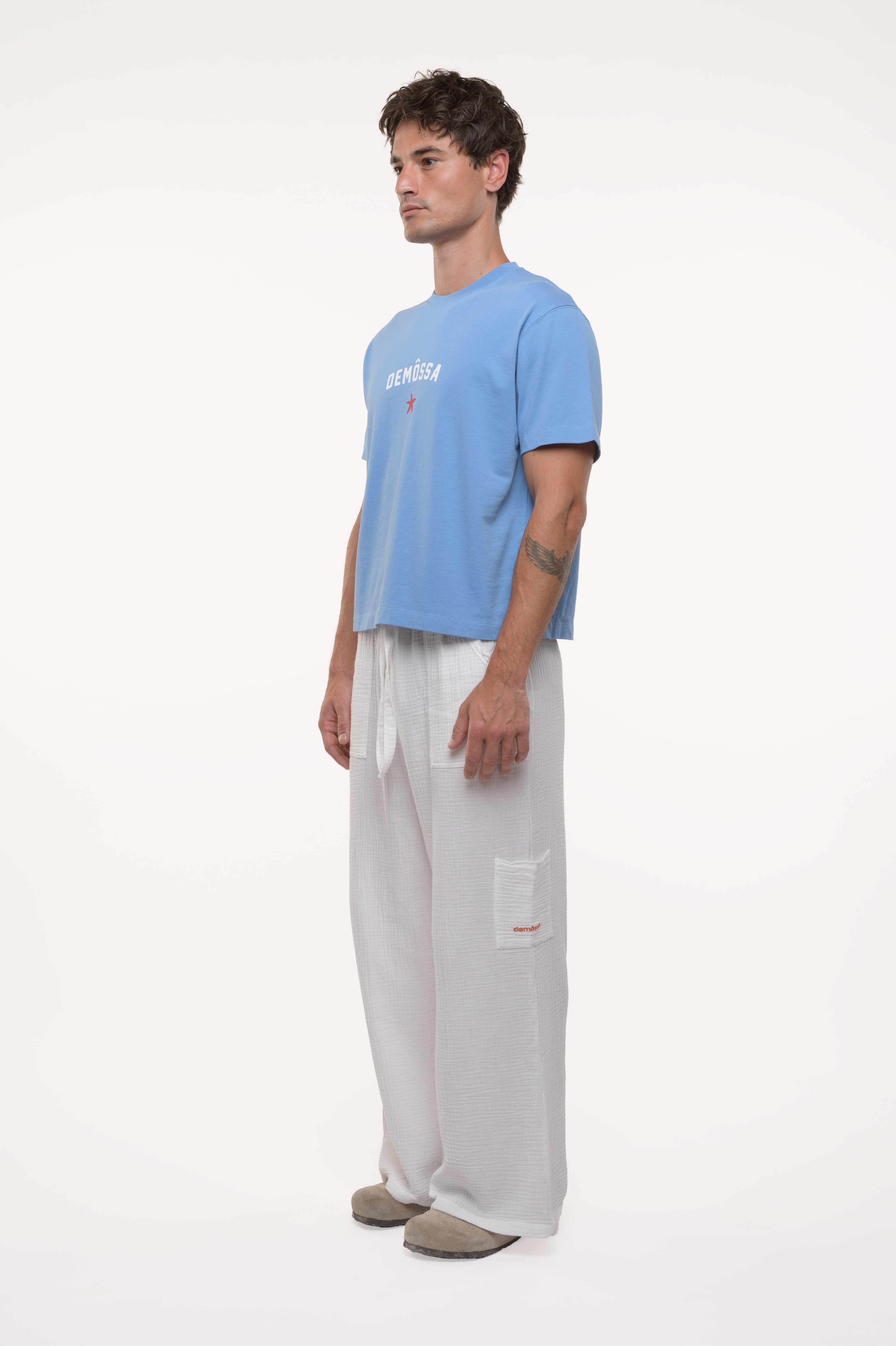 Side profile of a man wearing a light blue t-shirt and white pants on a white background
