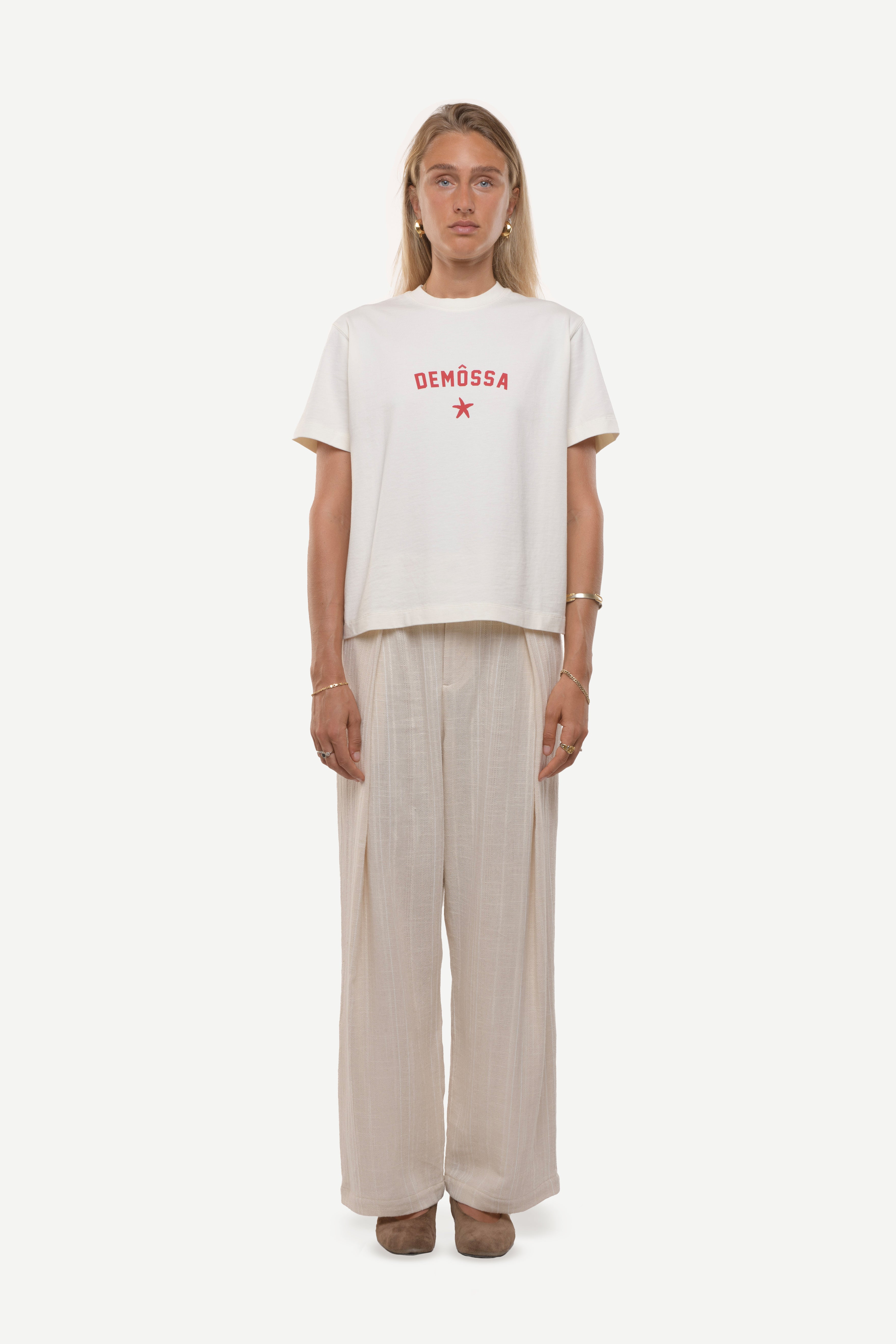 Woman wearing a white t-shirt with red demossa logo and beige pants on a white background