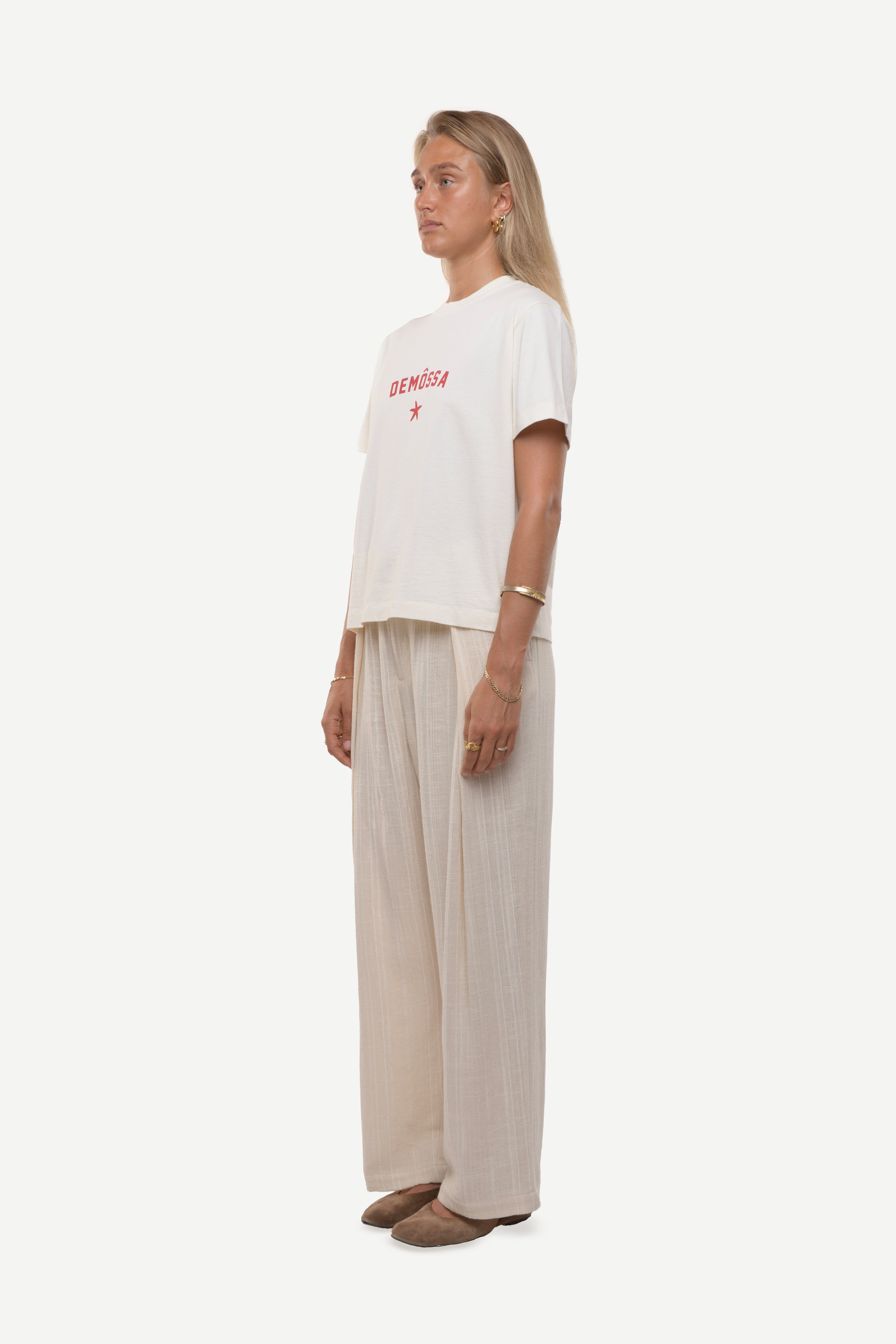 Side view of a woman wearing a white t-shirt with red demossa logo and beige pants on a white background