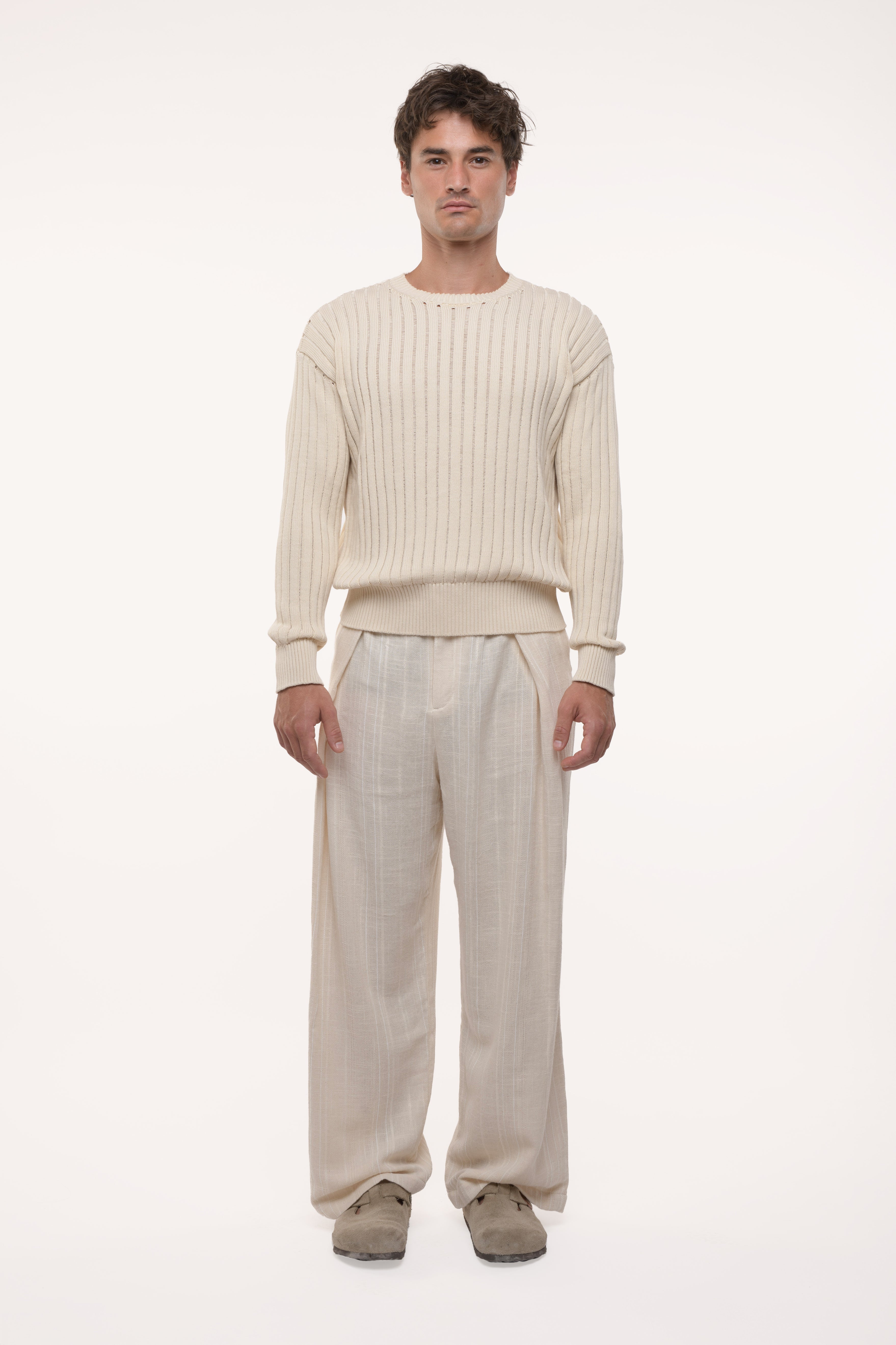 Man wearing a beige ribbed sweater and light-colored pants on a white background