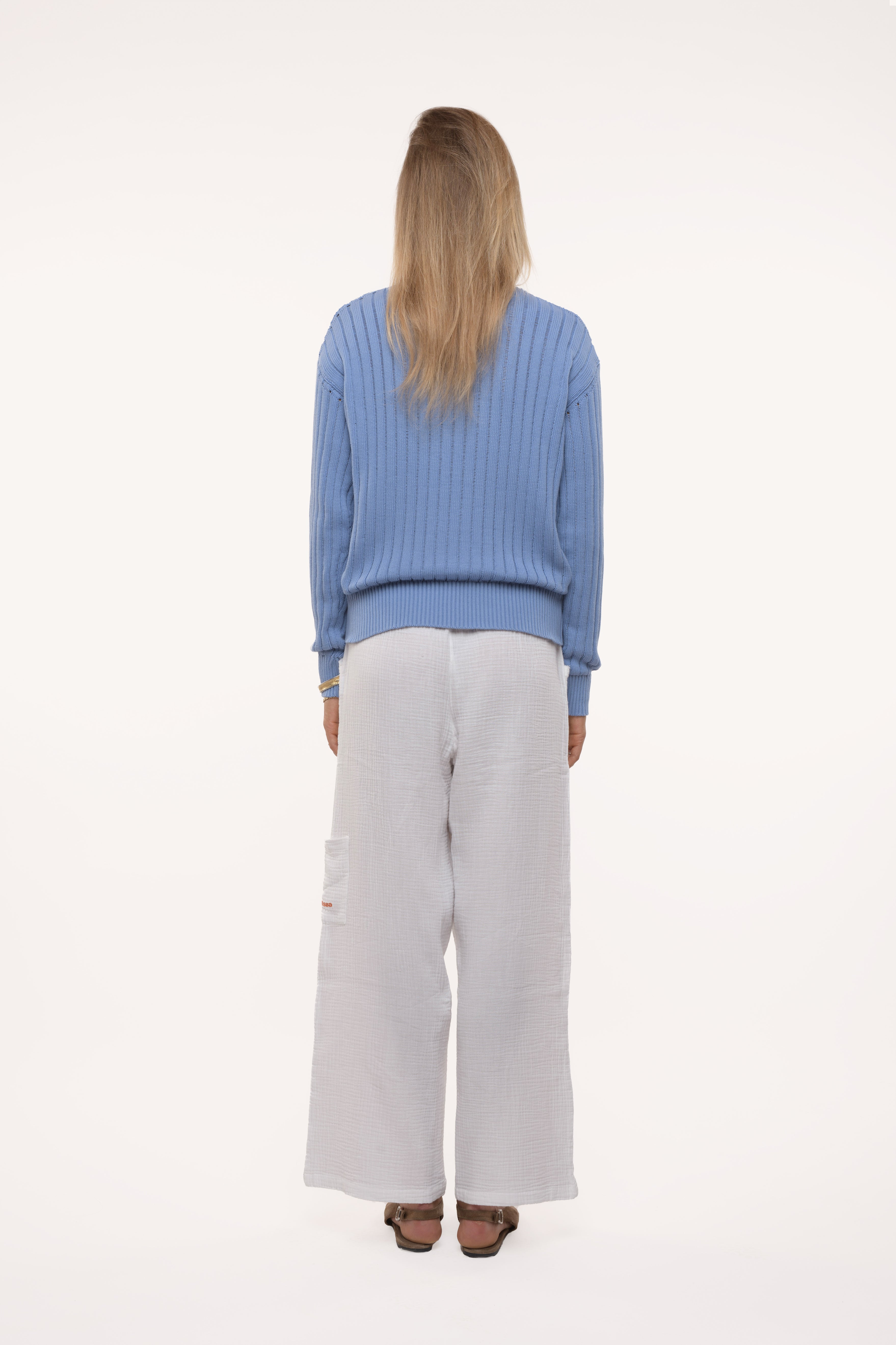 Back view of a woman wearing a blue sweater and light gray pants on a white background