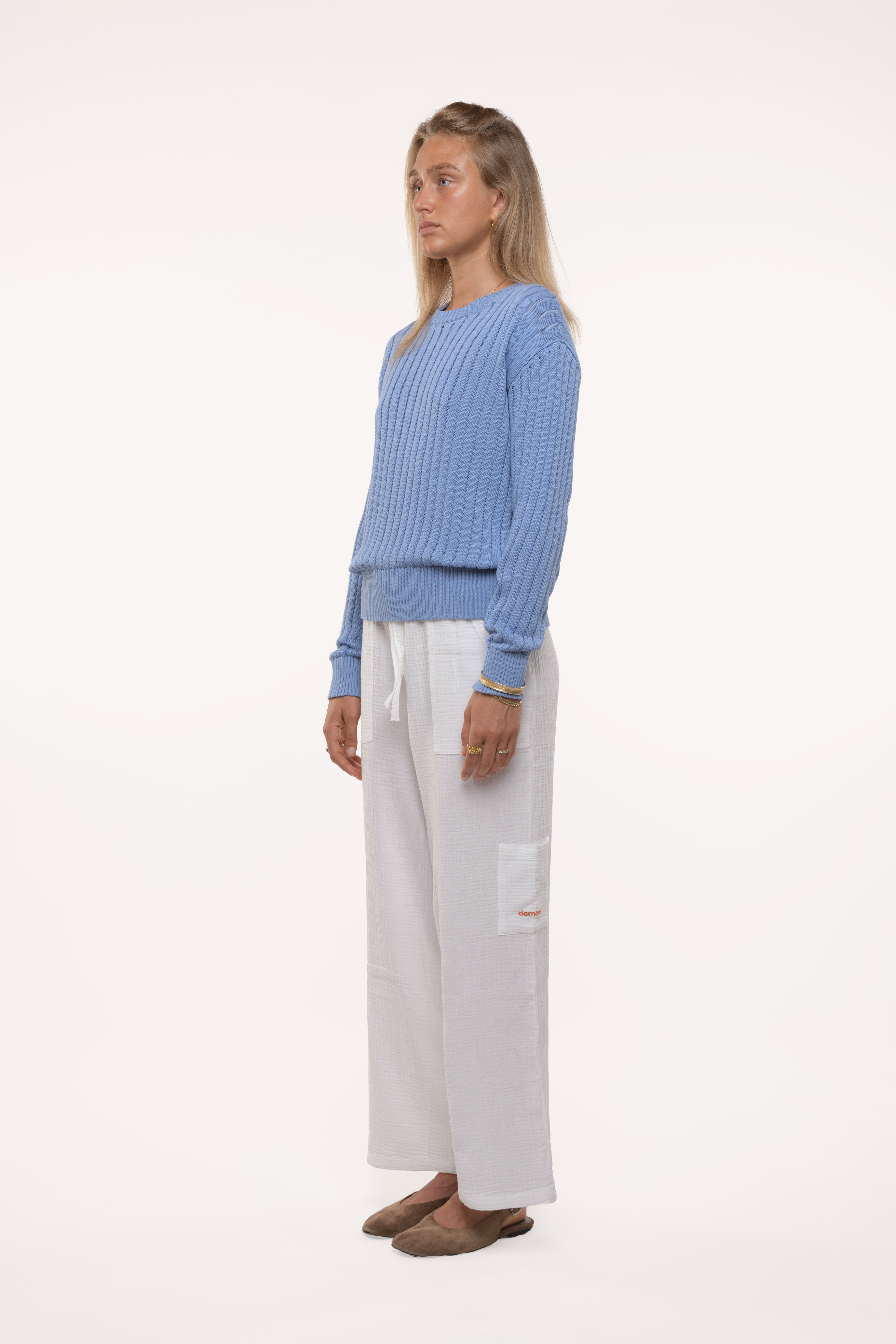 Side view of a woman wearing a blue open knit sweater and white pants on a white background