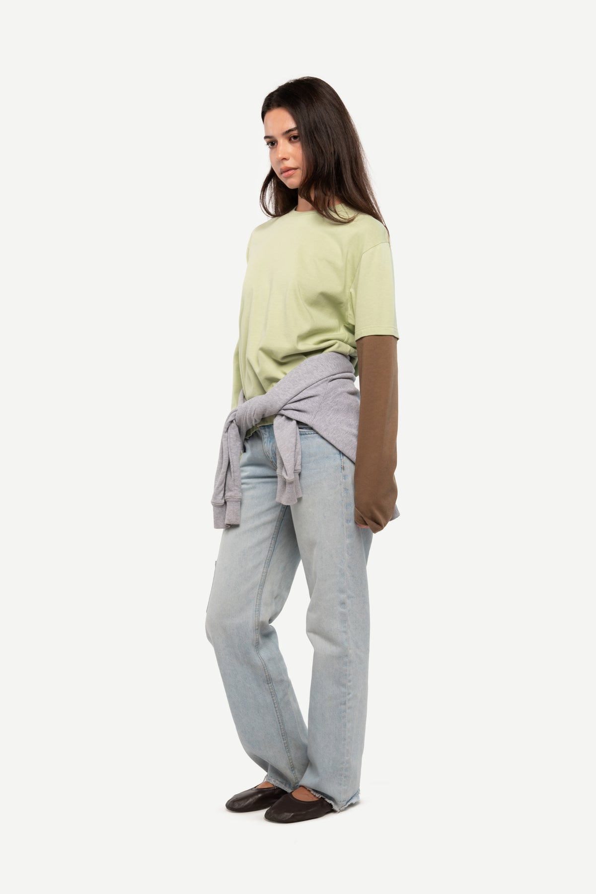 A woman modeling a light green short-sleeve top, light-wash wide-leg jeans, and a gray knit sweater tied around her waist, photographed against a plain light gray studio background.
