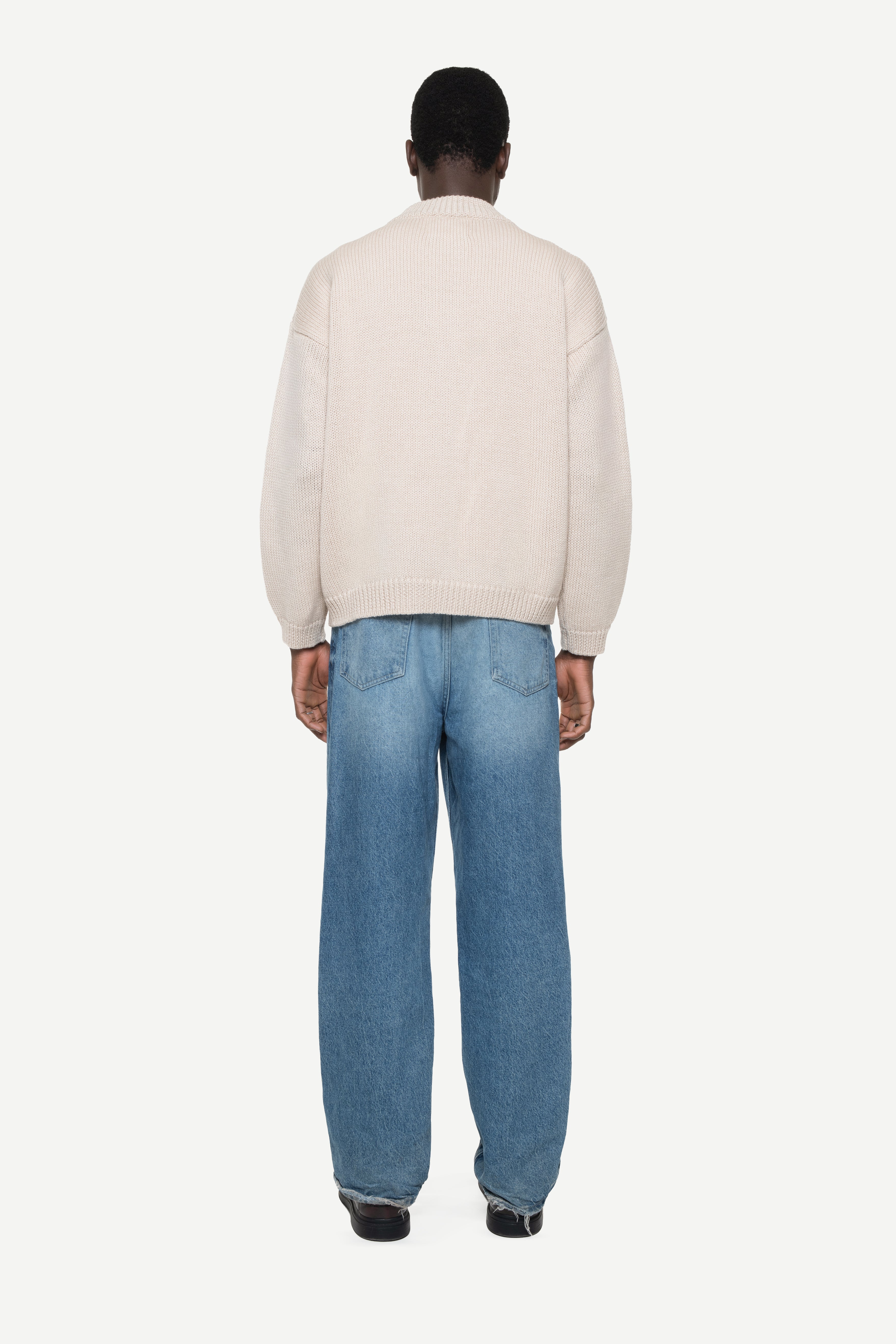 back view of a man wearing a beige buttoned cardigan and jeans on a white background