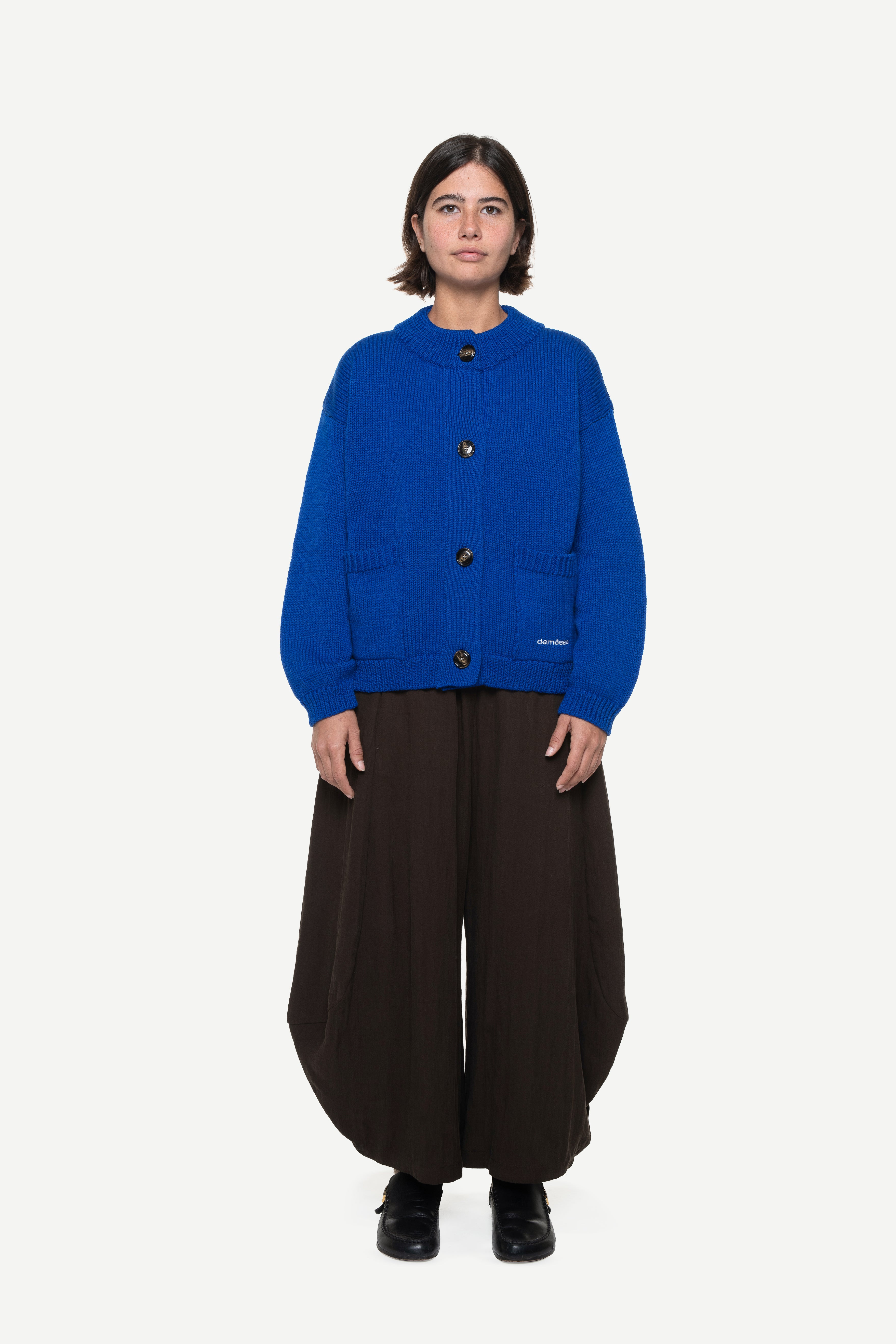 girl wearing a blue knit cardigan with two front pockets and brown pants on a white background
