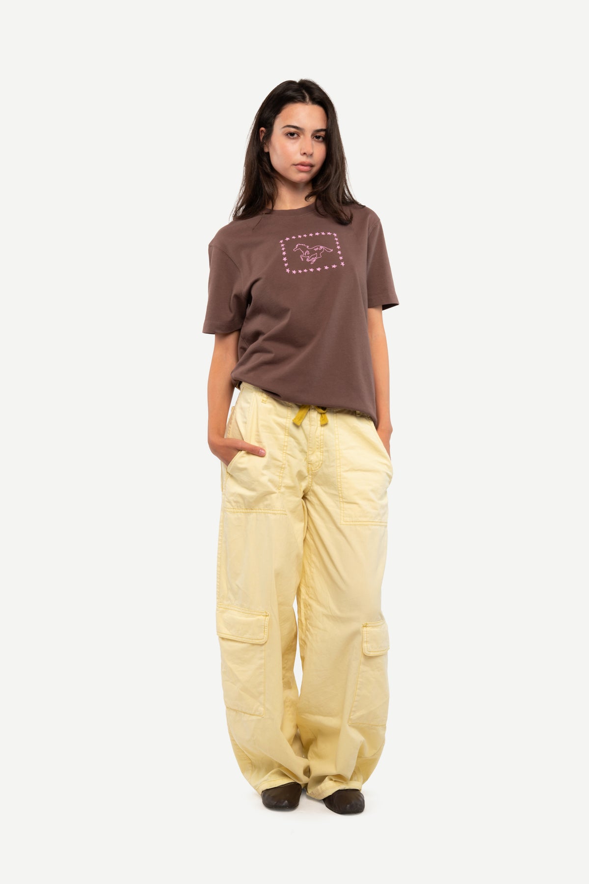 A model stands against a light gray background wearing a brown short-sleeve t-shirt with a graphic on the chest and loose, light yellow cargo pants.