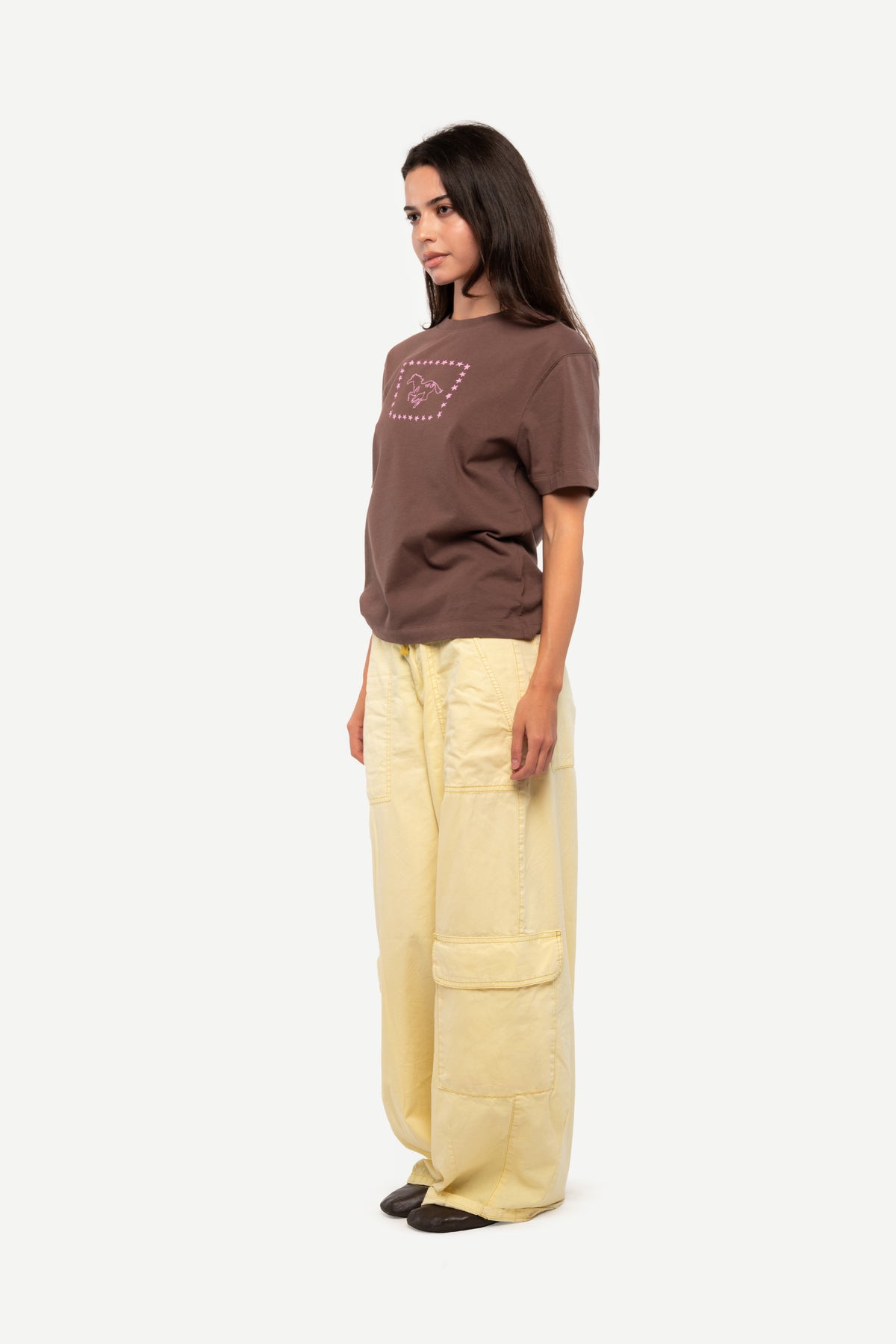 A woman modeling a brown short-sleeve t-shirt with a small graphic on the chest and wide-leg light yellow cargo pants, standing against a plain light gray background.