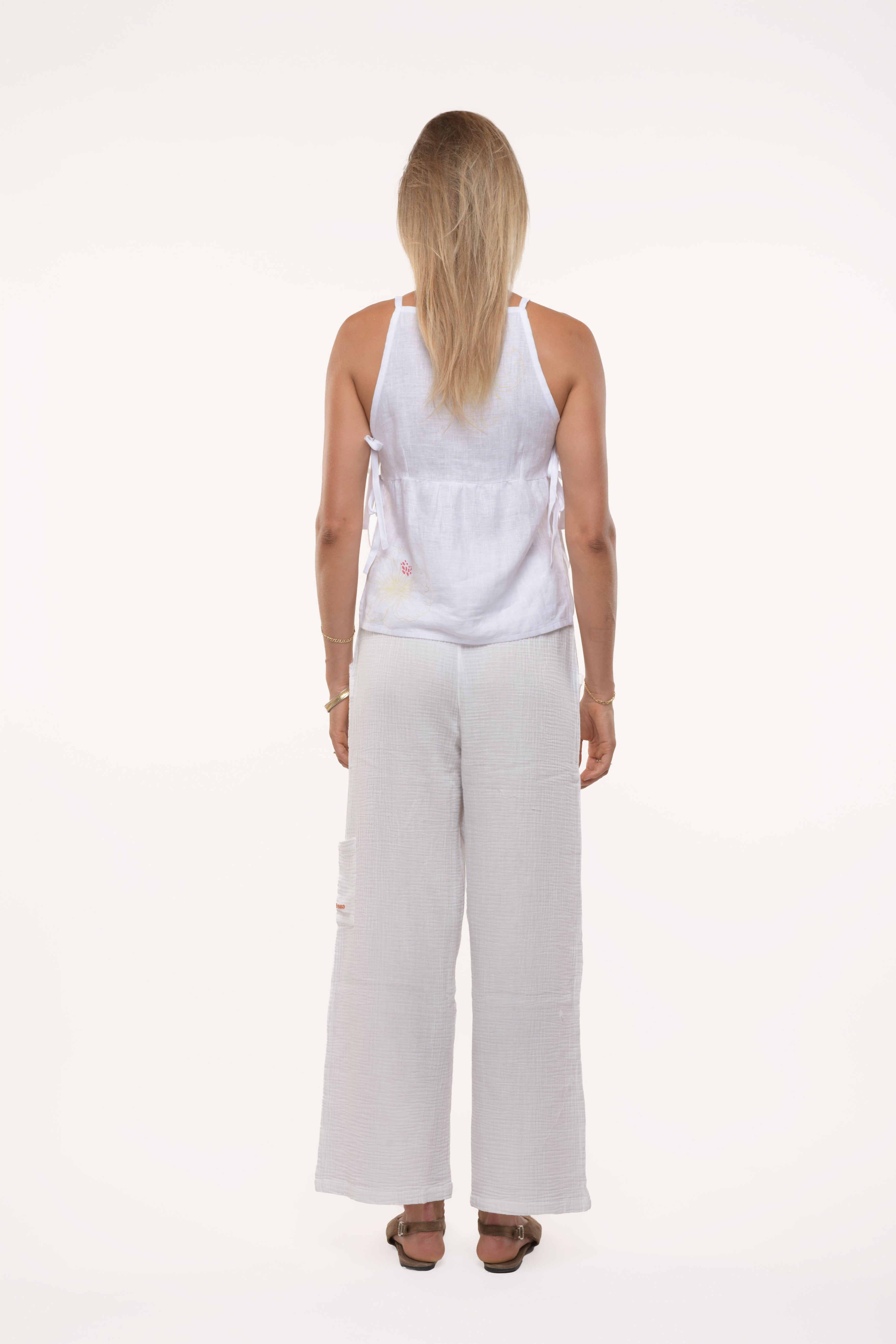 Back view of a woman wearing a white sleeveless linen top and white cotton pants on a white background