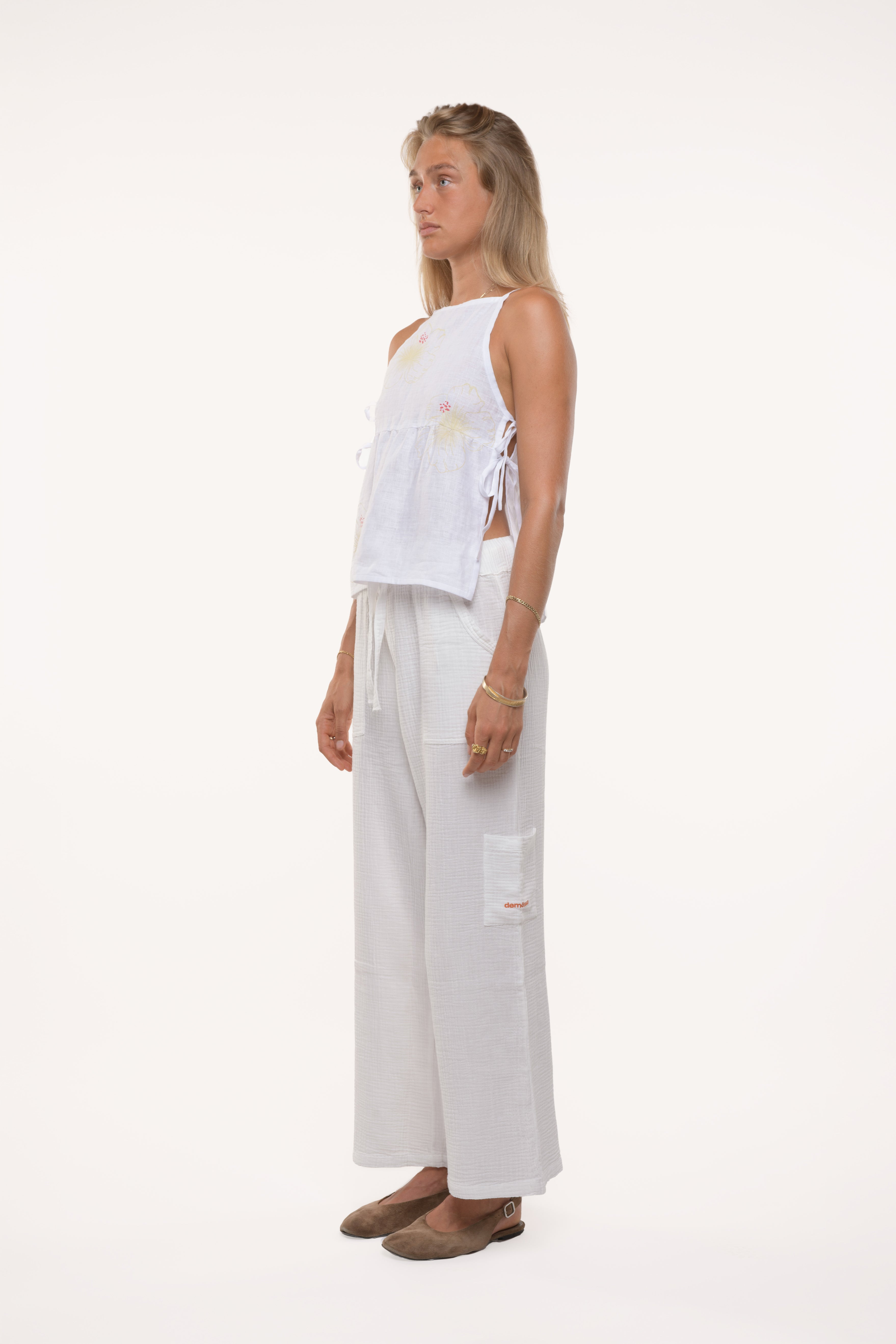 Side view of a woman wearing a white sleeveless top with adjustable ties and wide-leg pants on a white background