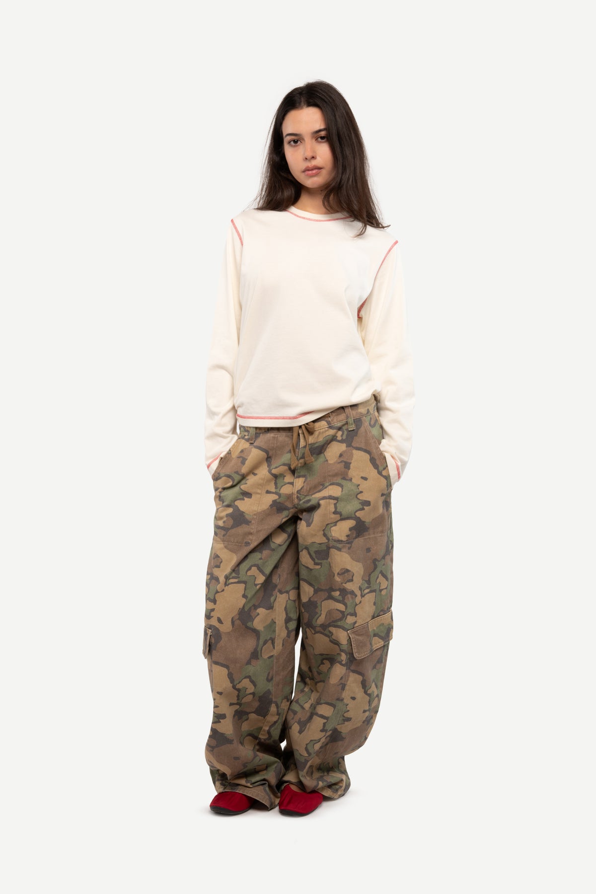 A model stands against a light gray background wearing a cream-colored long-sleeve top with red piping details, loose-fitting camouflage cargo pants, and red shoes.