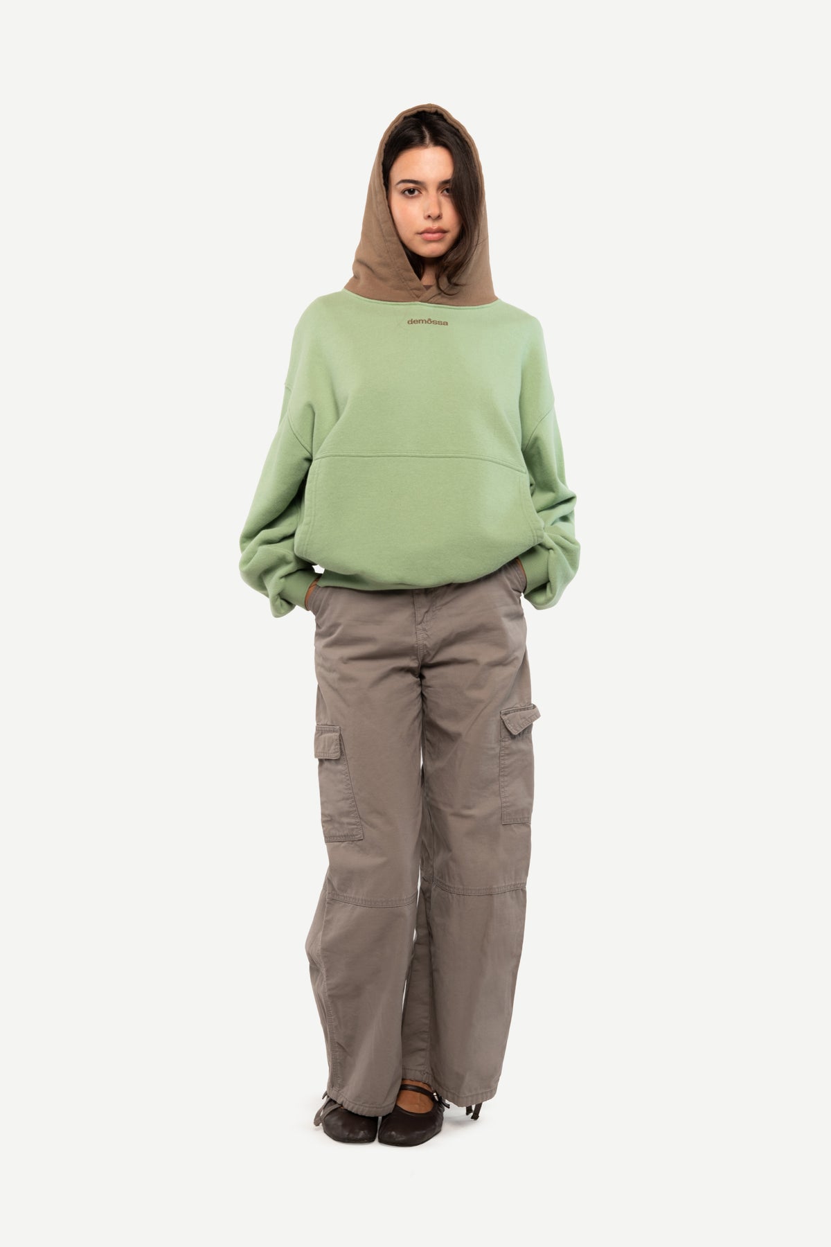 A model wearing a sage green oversized hoodie with a contrasting brown hood, paired with wide-leg taupe cargo pants and black shoes, against a plain light gray background.