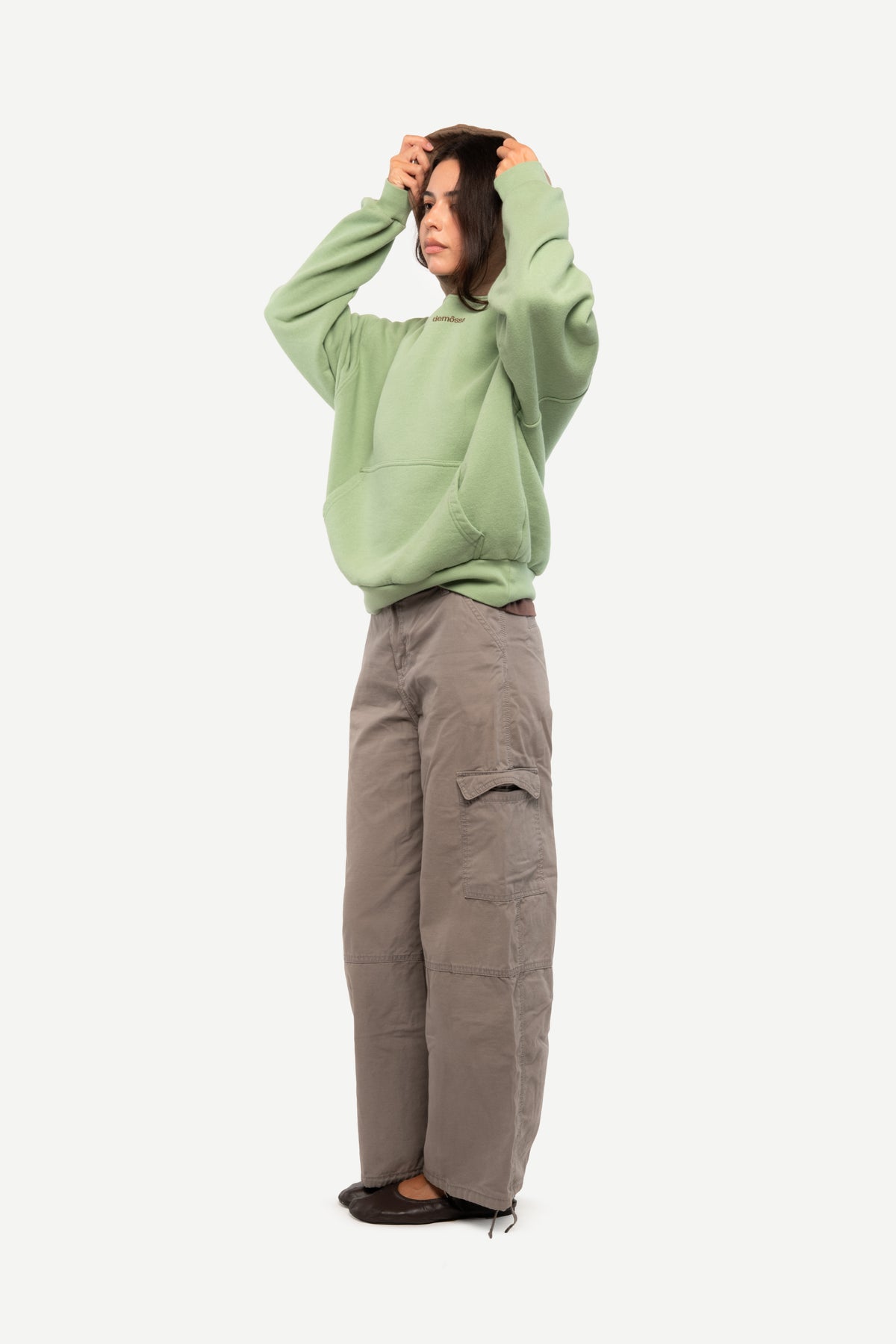 A model wearing a sage green oversized hoodie and wide-leg taupe cargo pants, standing against a plain light gray background.