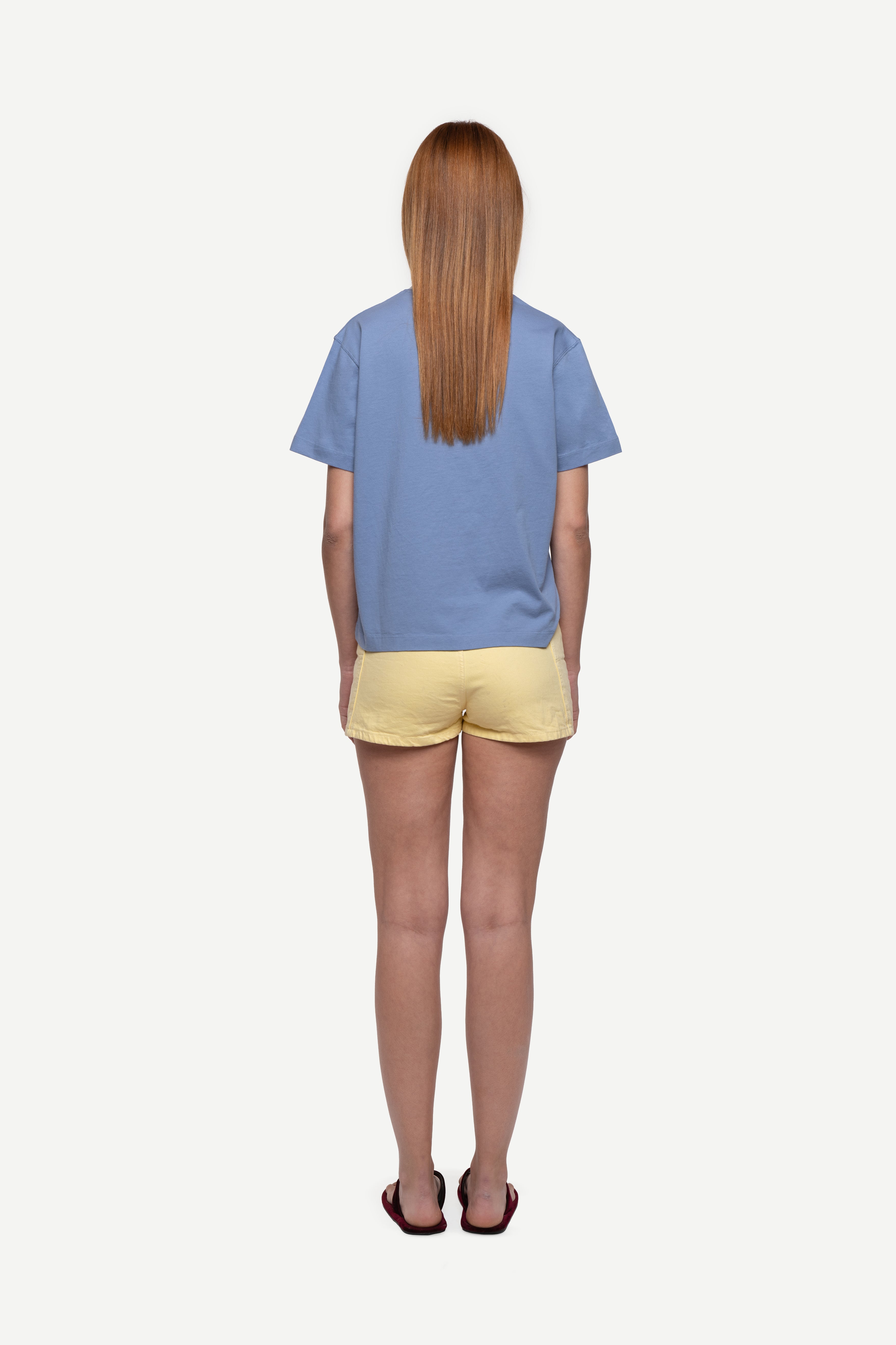 back view of woman wearing a regular fit blue short-sleeved t-shirt, yellow shorts and flip flops on a white background