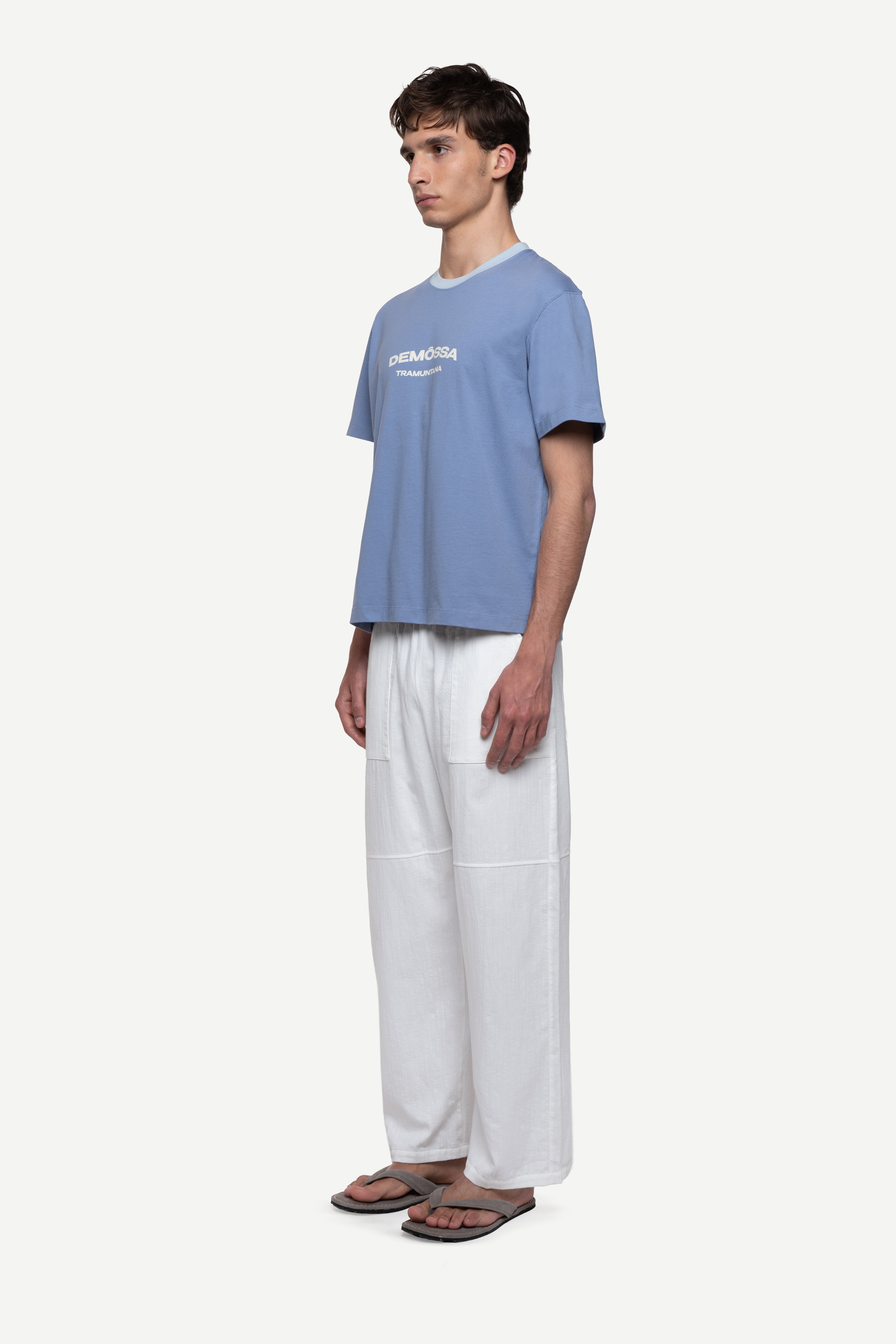 side view of man wearing a regular fit blue short-sleeved t-shirt with ivory demôssa print on the chest, white summer trousers and flip flops on a white background
