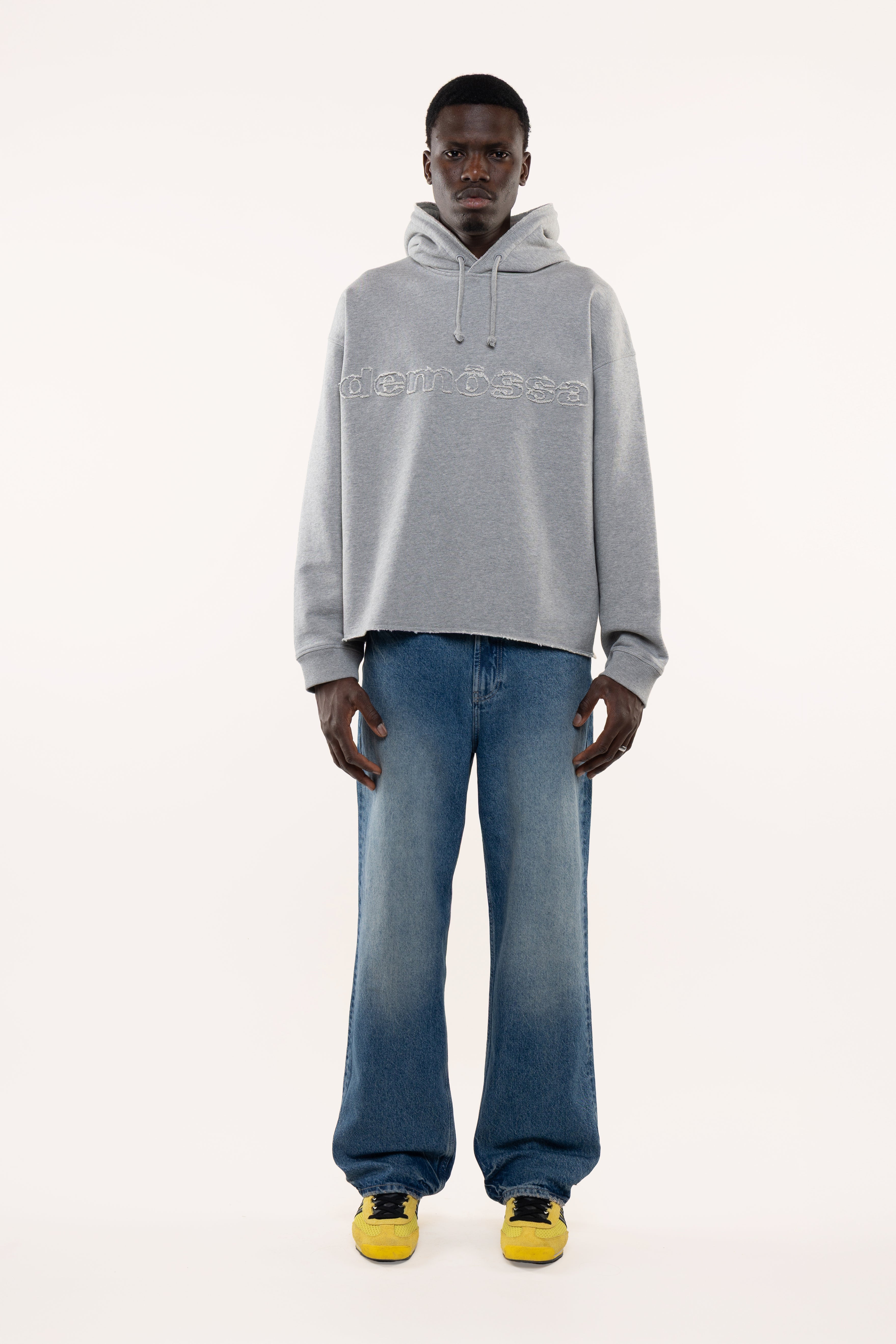 Man wearing a gray hoodie with demossa branding and blue jeans on a white background