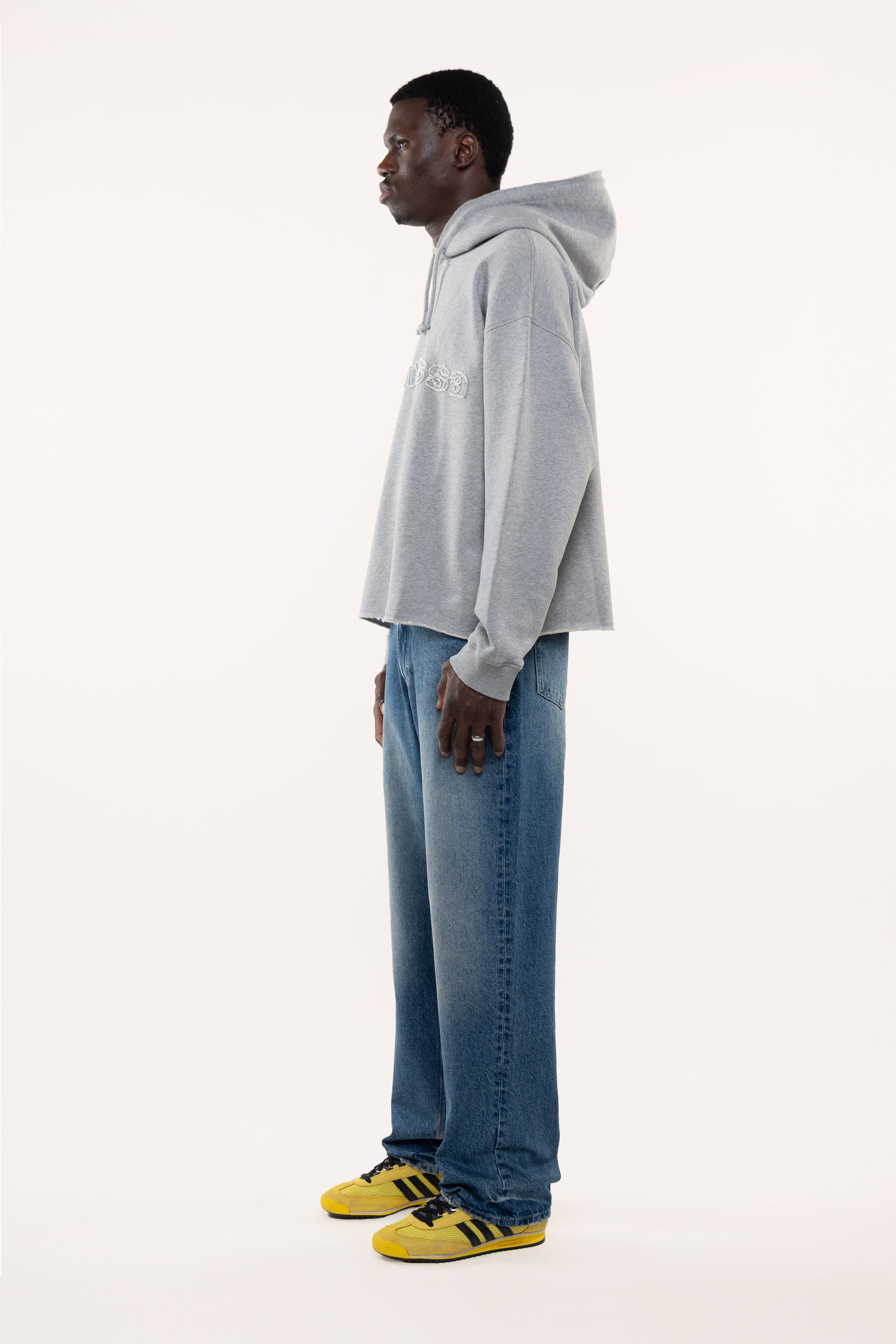 Side view of a man wearing a gray hoodie and blue jeans standing against a white background