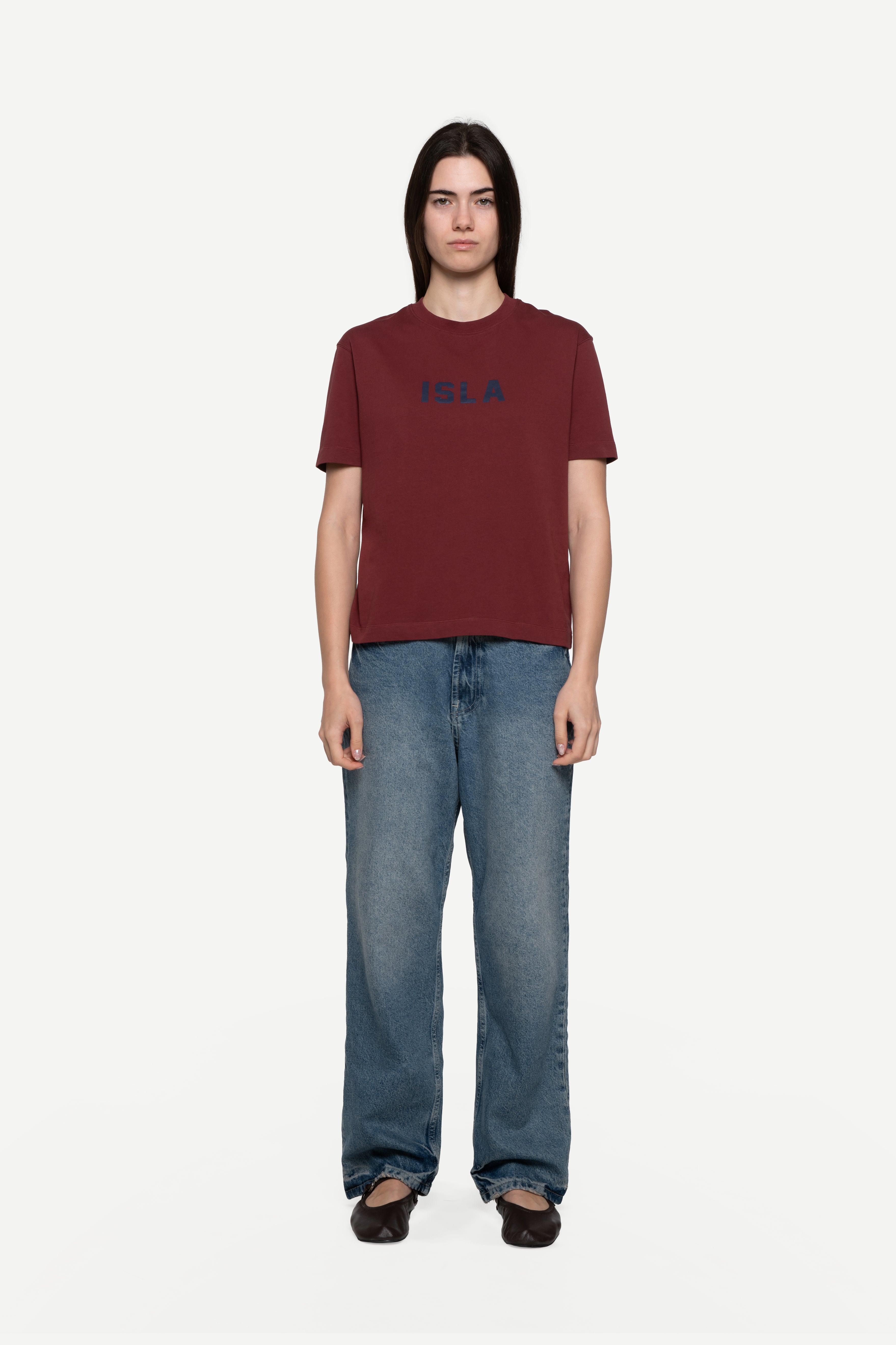 front view of woman wearing a regular fit short-sleeved burgundy t-shirt featuring a tonal 'demôssa' logo print on the chest with blue jeans on a white background