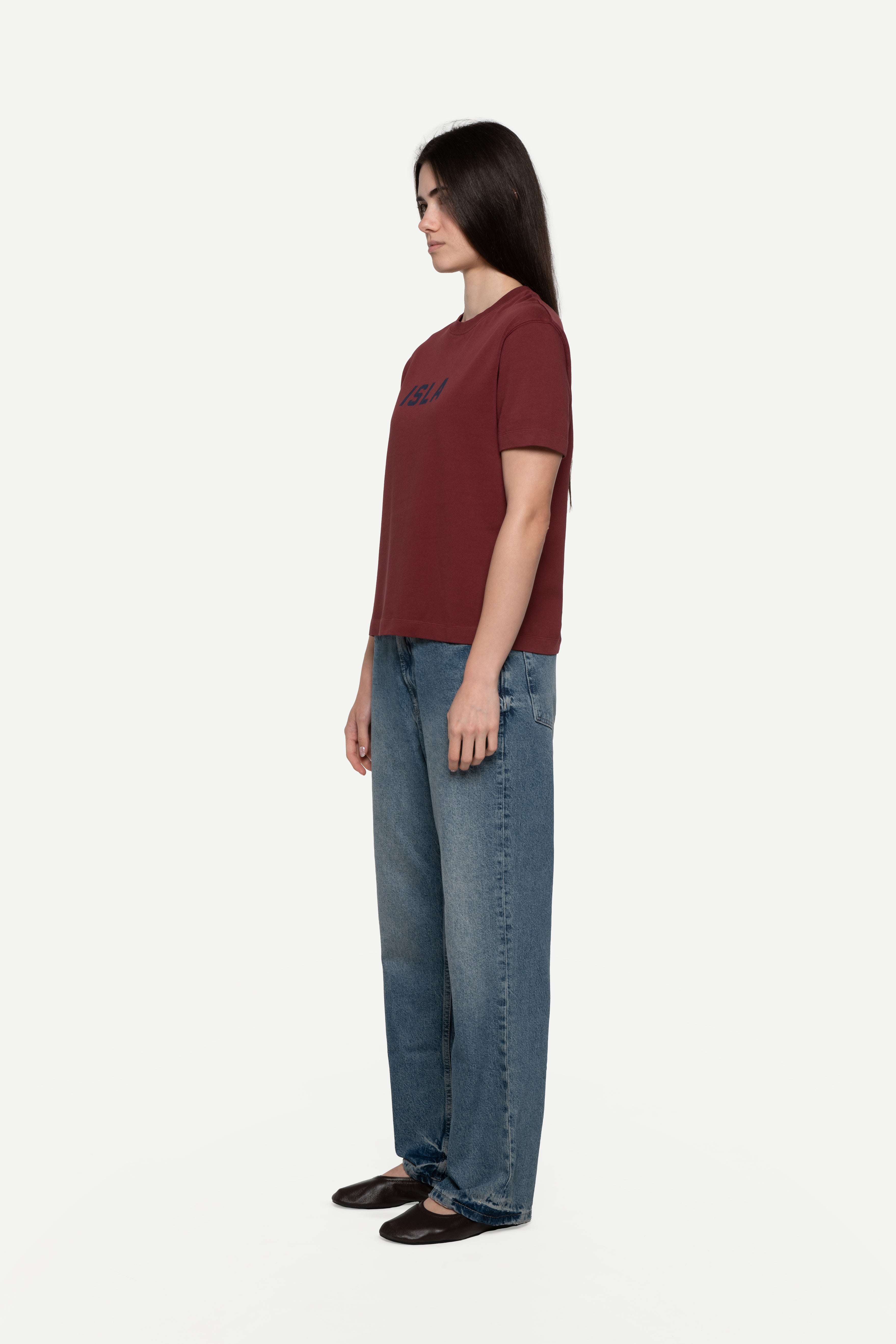 side view of woman wearing a regular fit short-sleeved burgundy t-shirt featuring a tonal 'demôssa' logo print on the chest with blue jeans on a white background