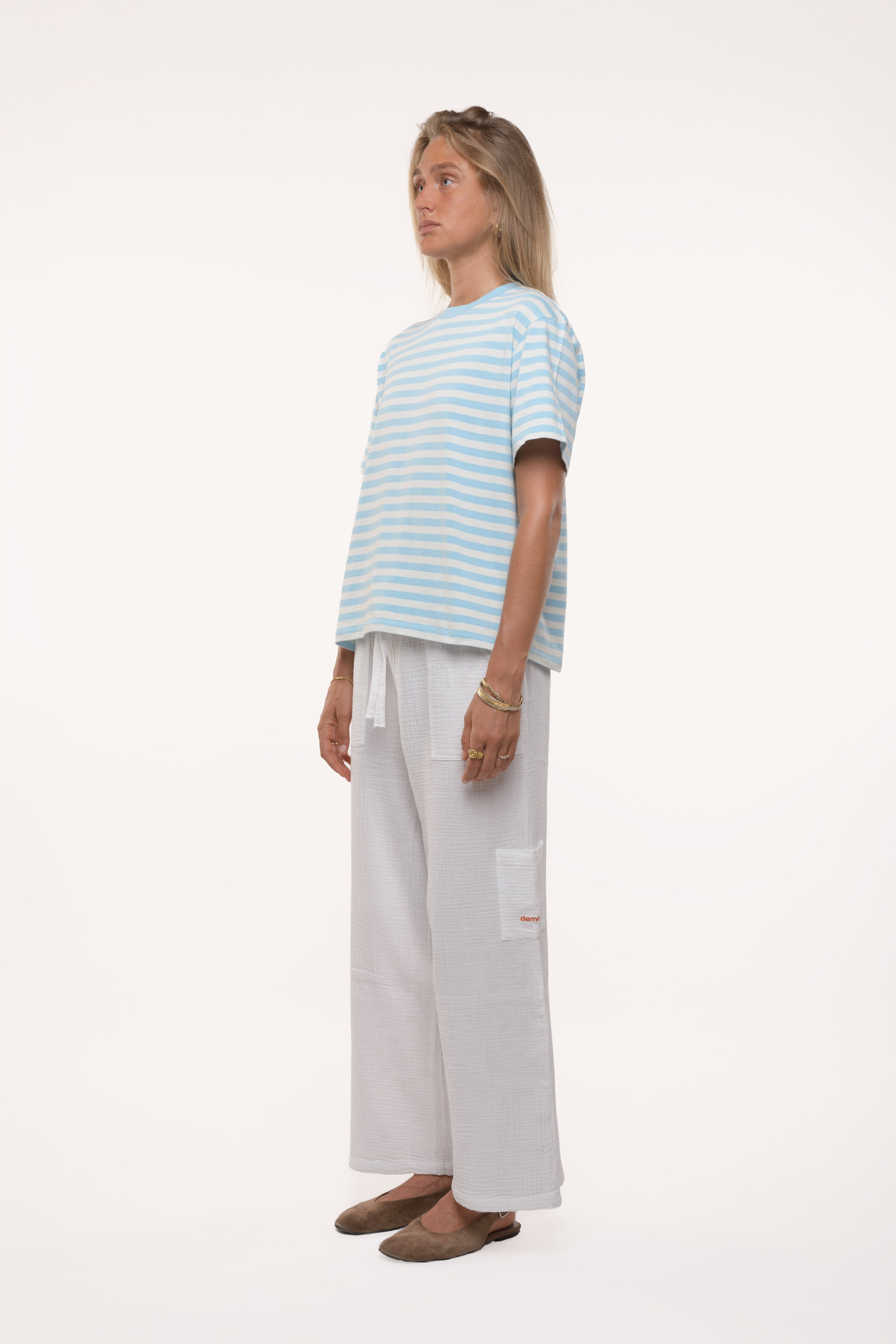 Side view of a woman wearing a light blue striped shirt and white pants on a white background