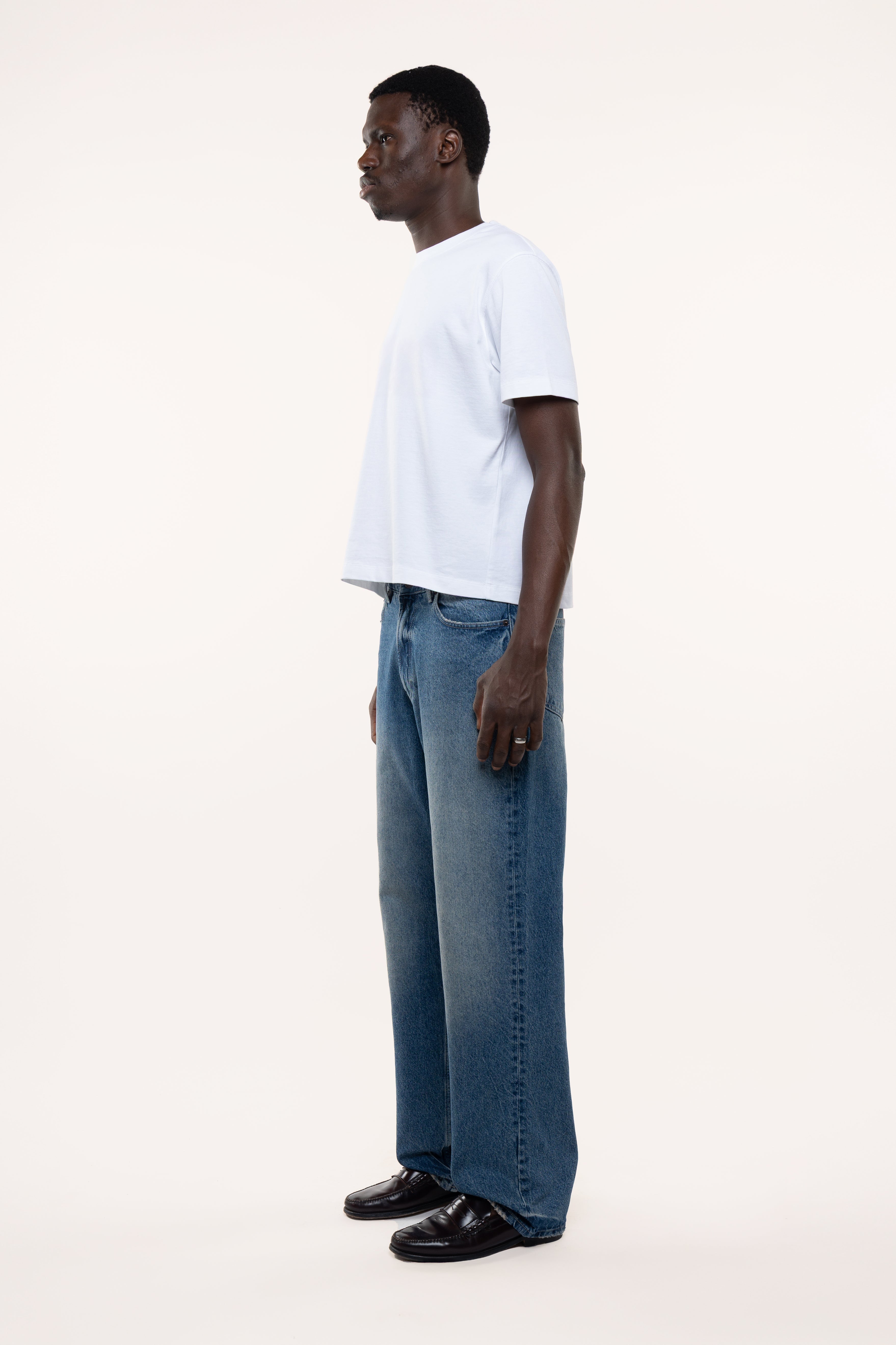Side view of a man wearing a white t-shirt and blue jeans standing against a plain background