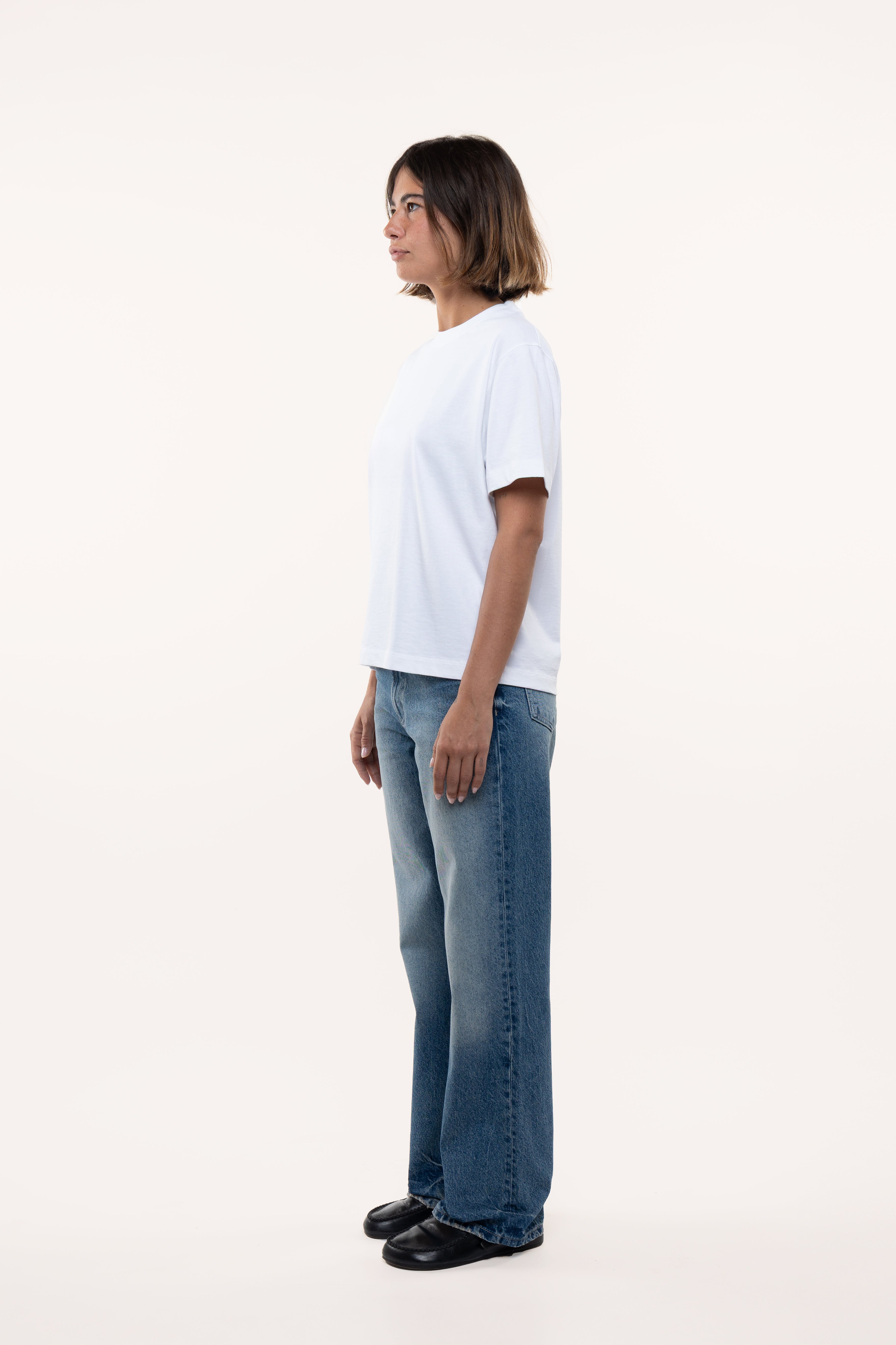 Side view of a woman wearing a white t-shirt and blue jeans standing against a white background