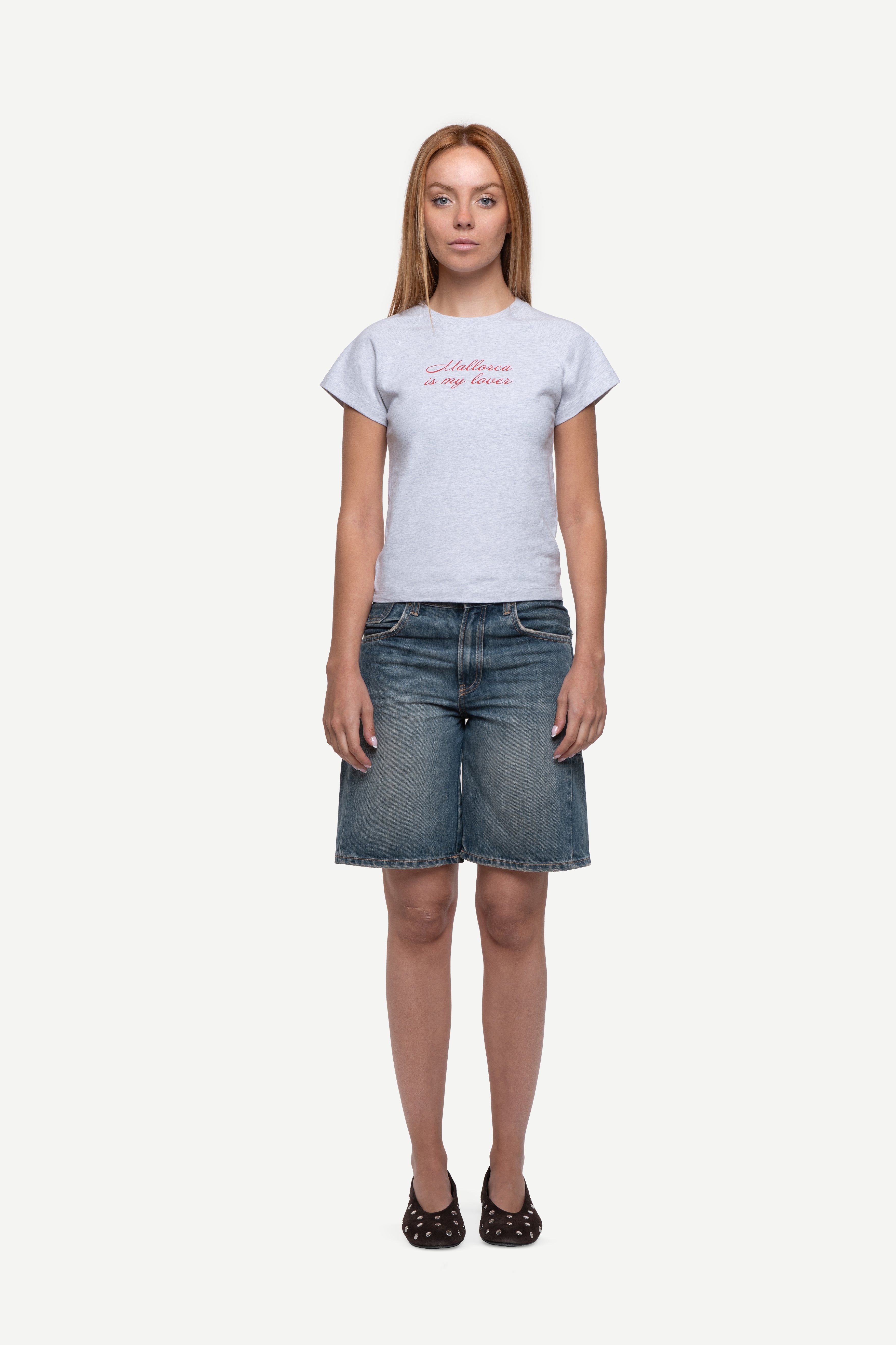 front view of woman wearing a fitted short-sleeved t-shirt with "Mallorca is my lover" front print in bright red, blue short jeans and ballet flats on a white background