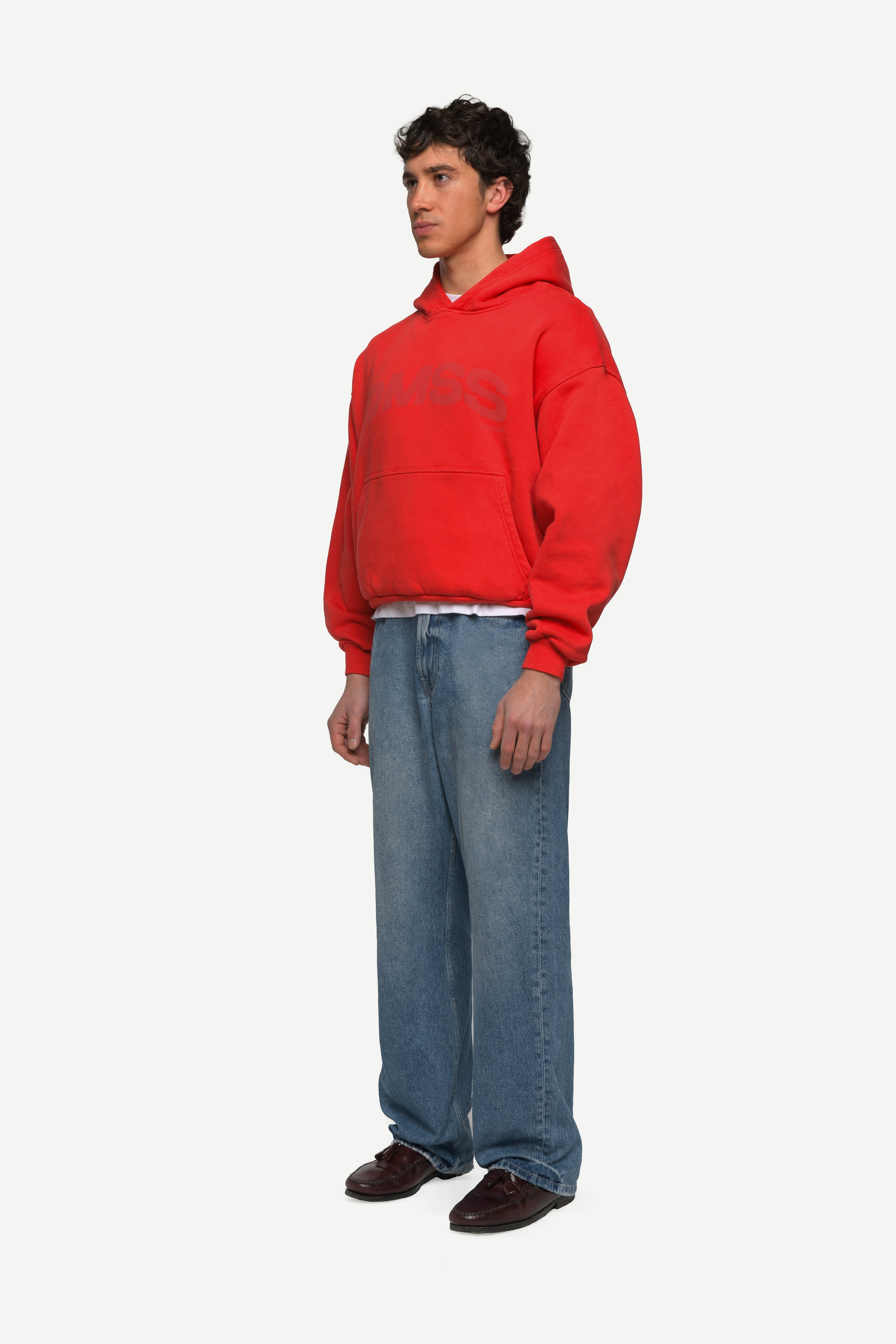 side view of man wearing a red hoodie with large DMSS logo in a subtle tone and blue jeans on a white background