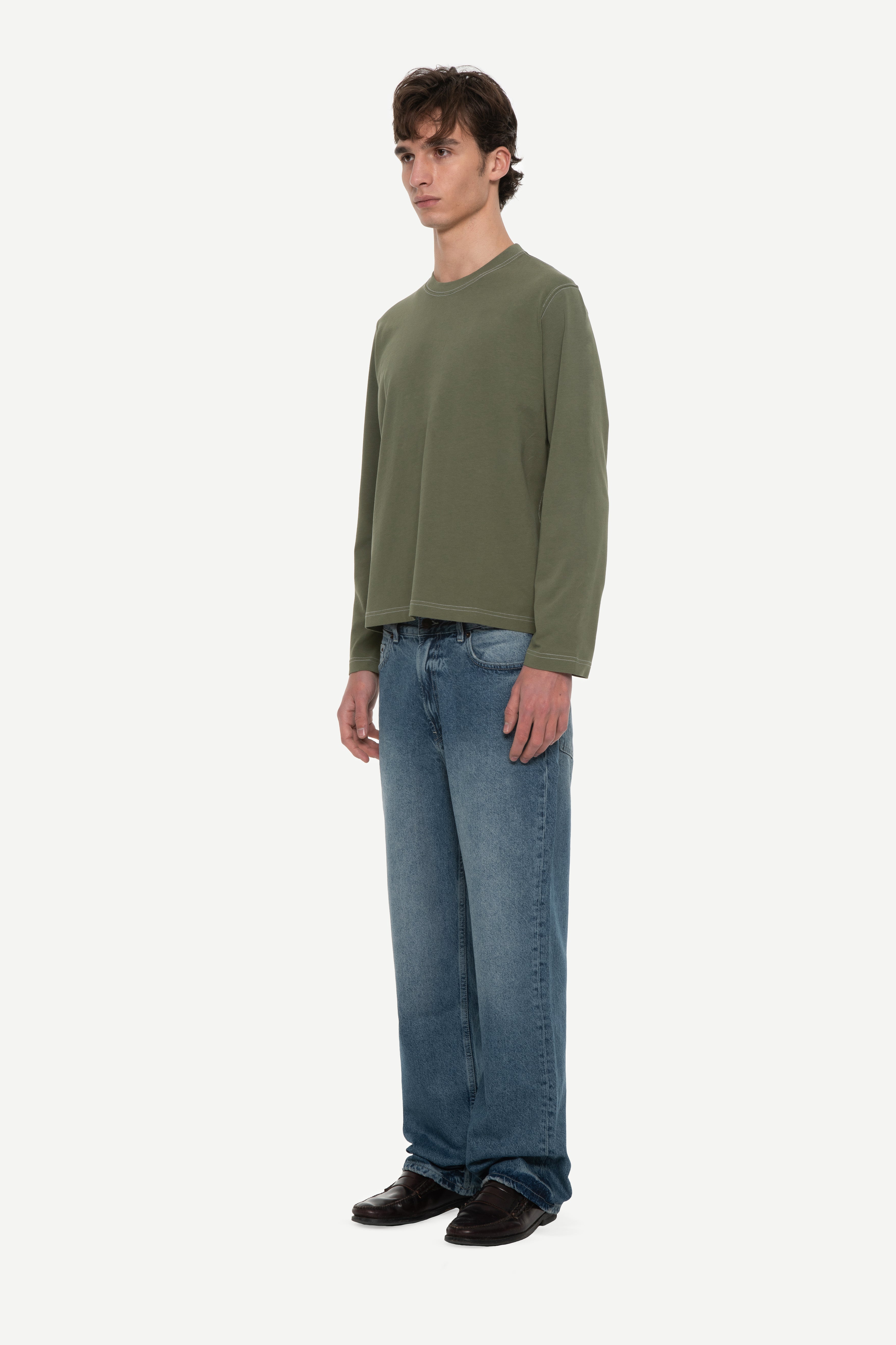 side view of man wearing a military green long sleeve tee with contrast stitching and blue jeans on a white background
