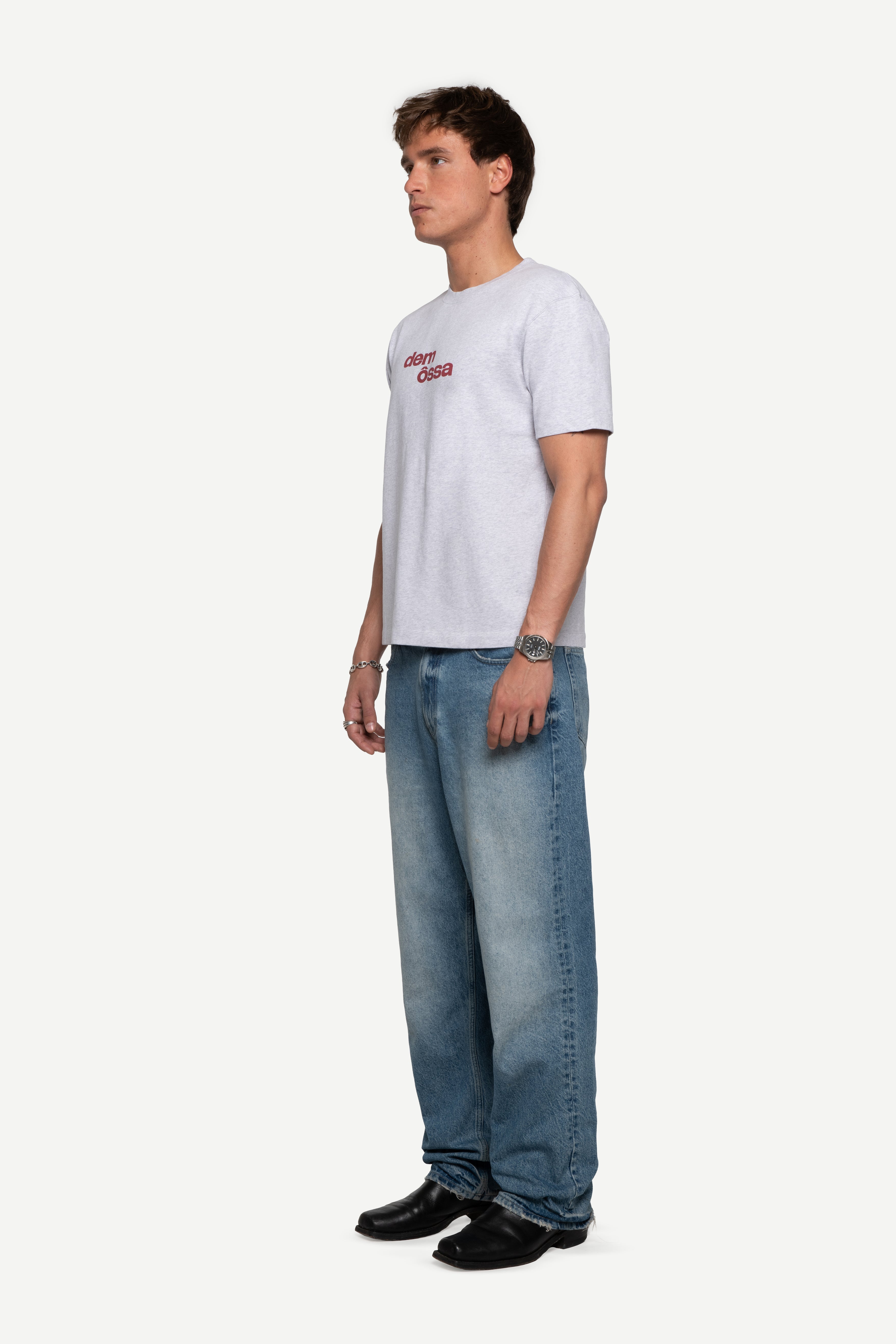 side view of man wearing a regular fit short-sleeved grey melange t-shirt featuring a tonal 'demôssa' logo print on the chest with blue jeans on a white background