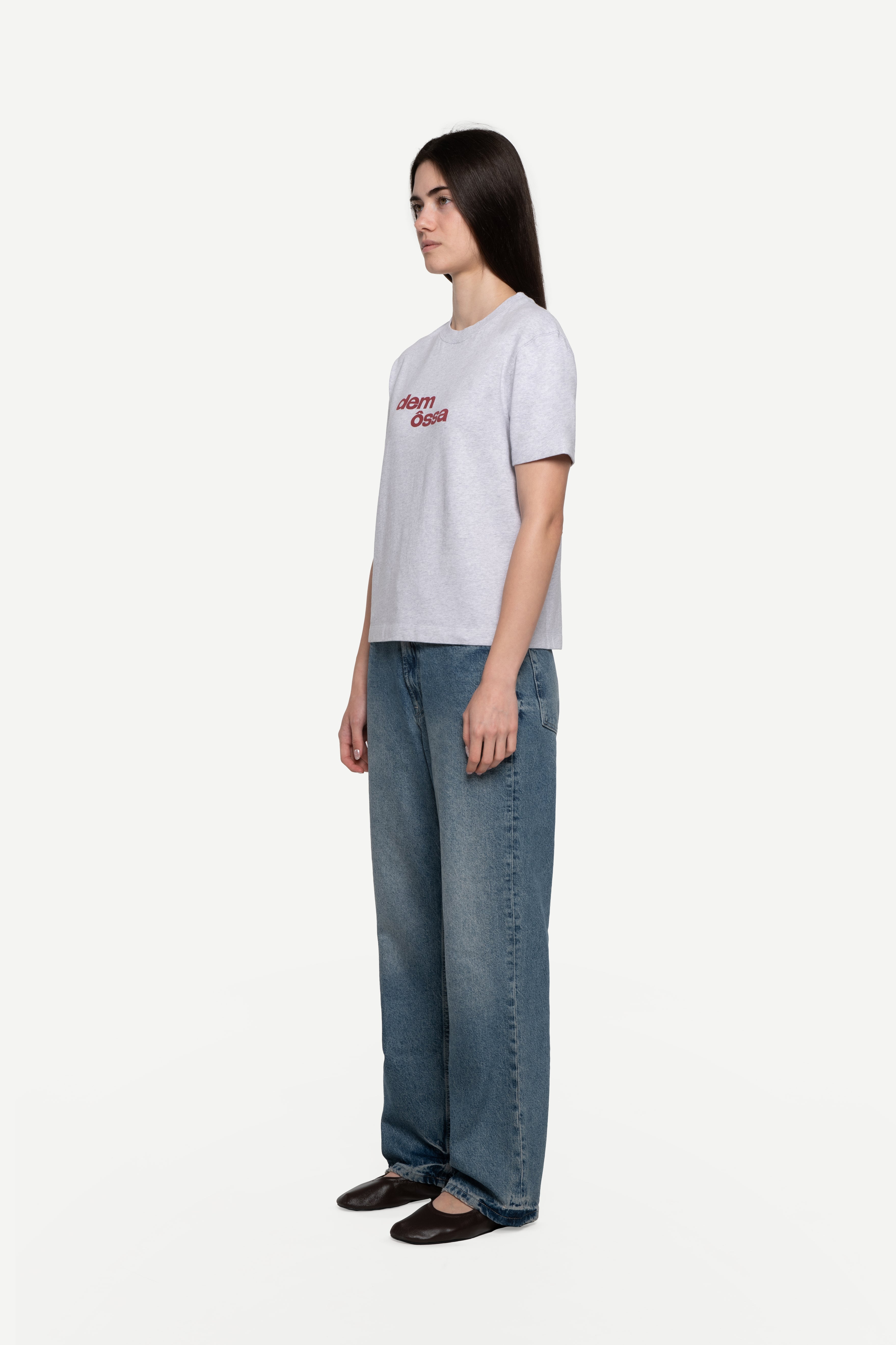 side view of woman wearing a regular fit short-sleeved grey melange t-shirt featuring a tonal 'demôssa' logo print on the chest with blue jeans on a white background