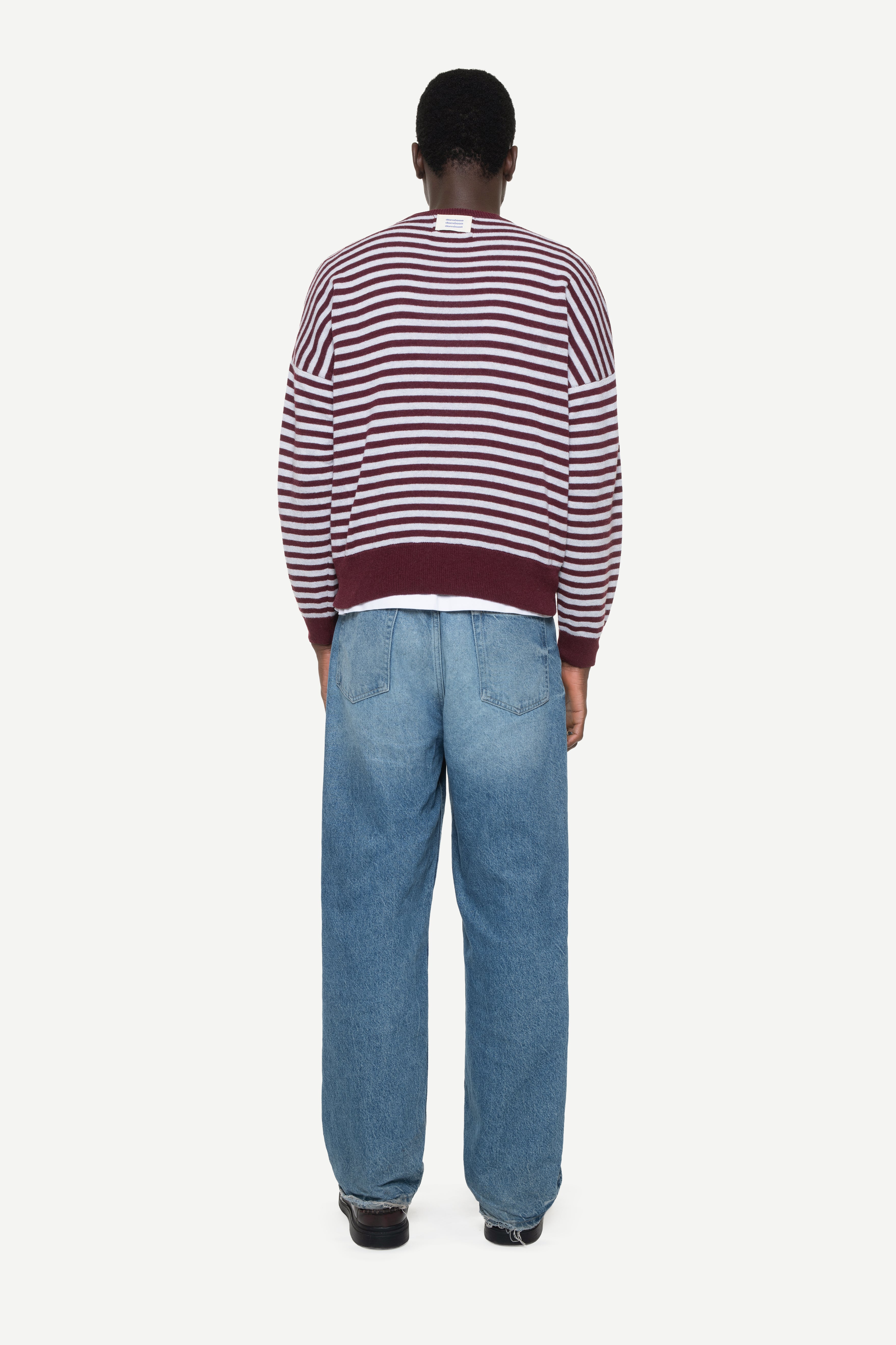 back view of a man wearing a burgundy knit striped sweater with demossa label and jeans on a white background