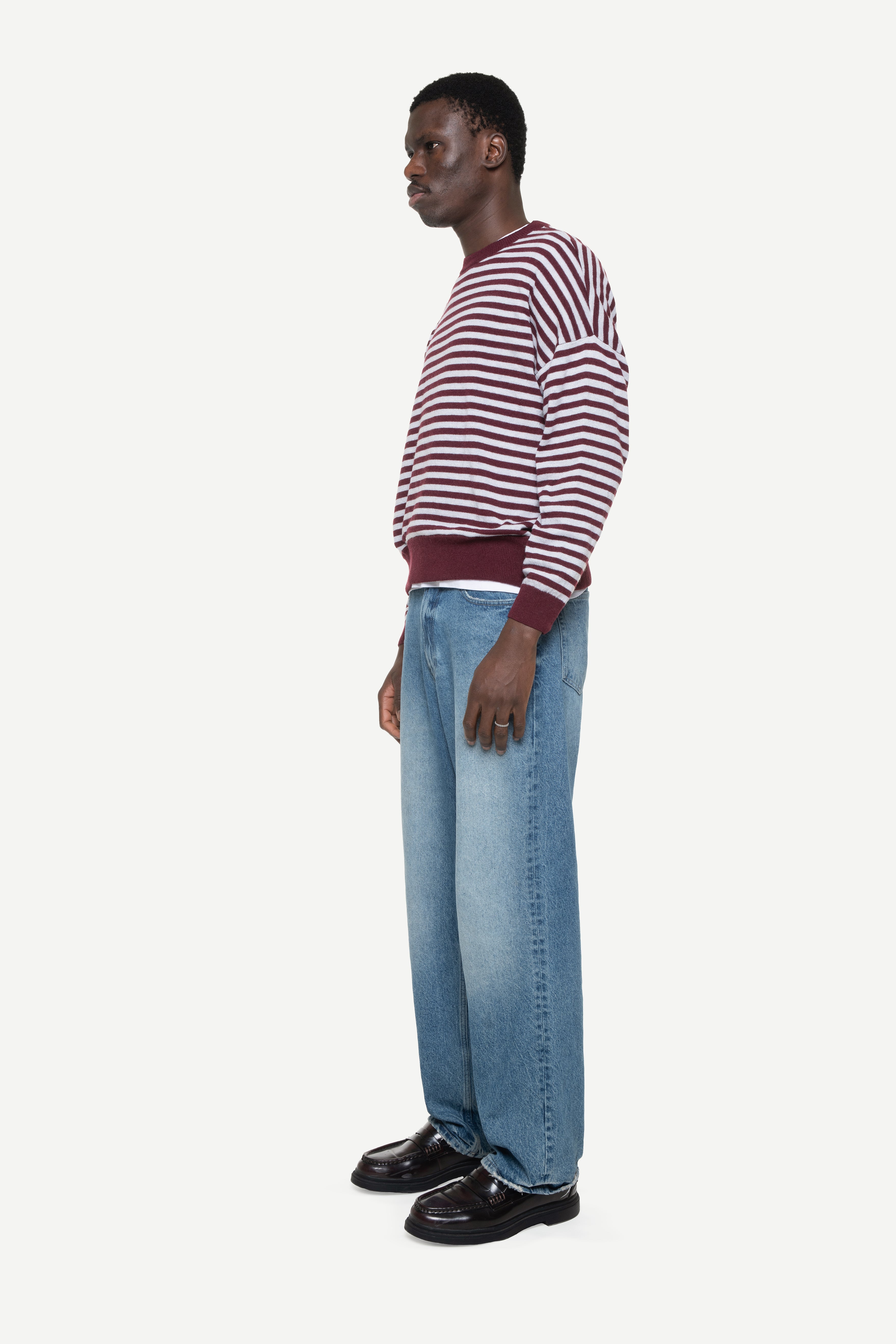 side view of a man wearing a burgundy knit striped sweater and jeans on a white background