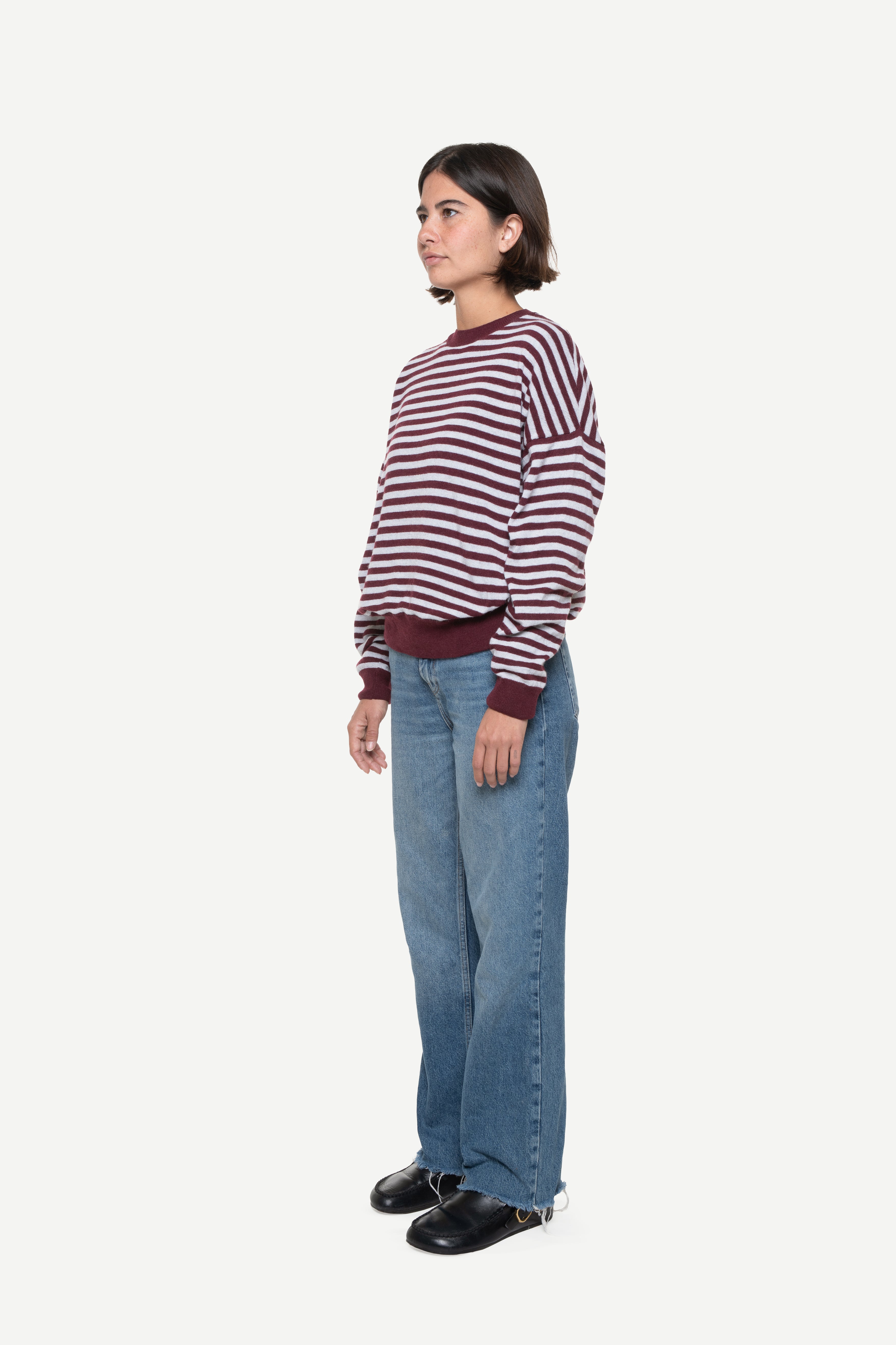 side view of a woman wearing a burgundy knit striped sweater and jeans on a white background