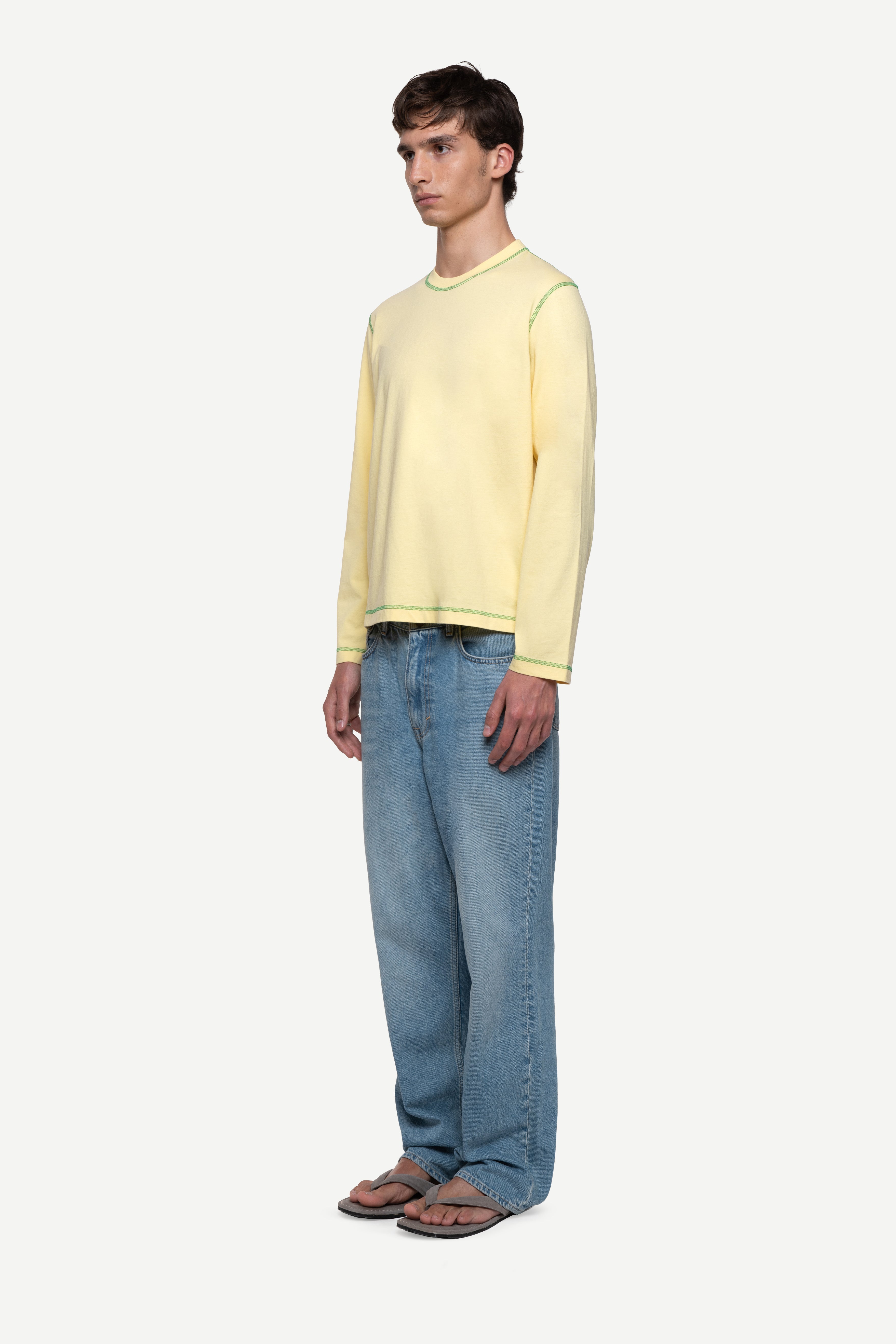 side view of man wearing a regular fit pale yellow red long-sleeved t-shirt with contrast green over-lock stitching, blue jeans and flip flops on a white background