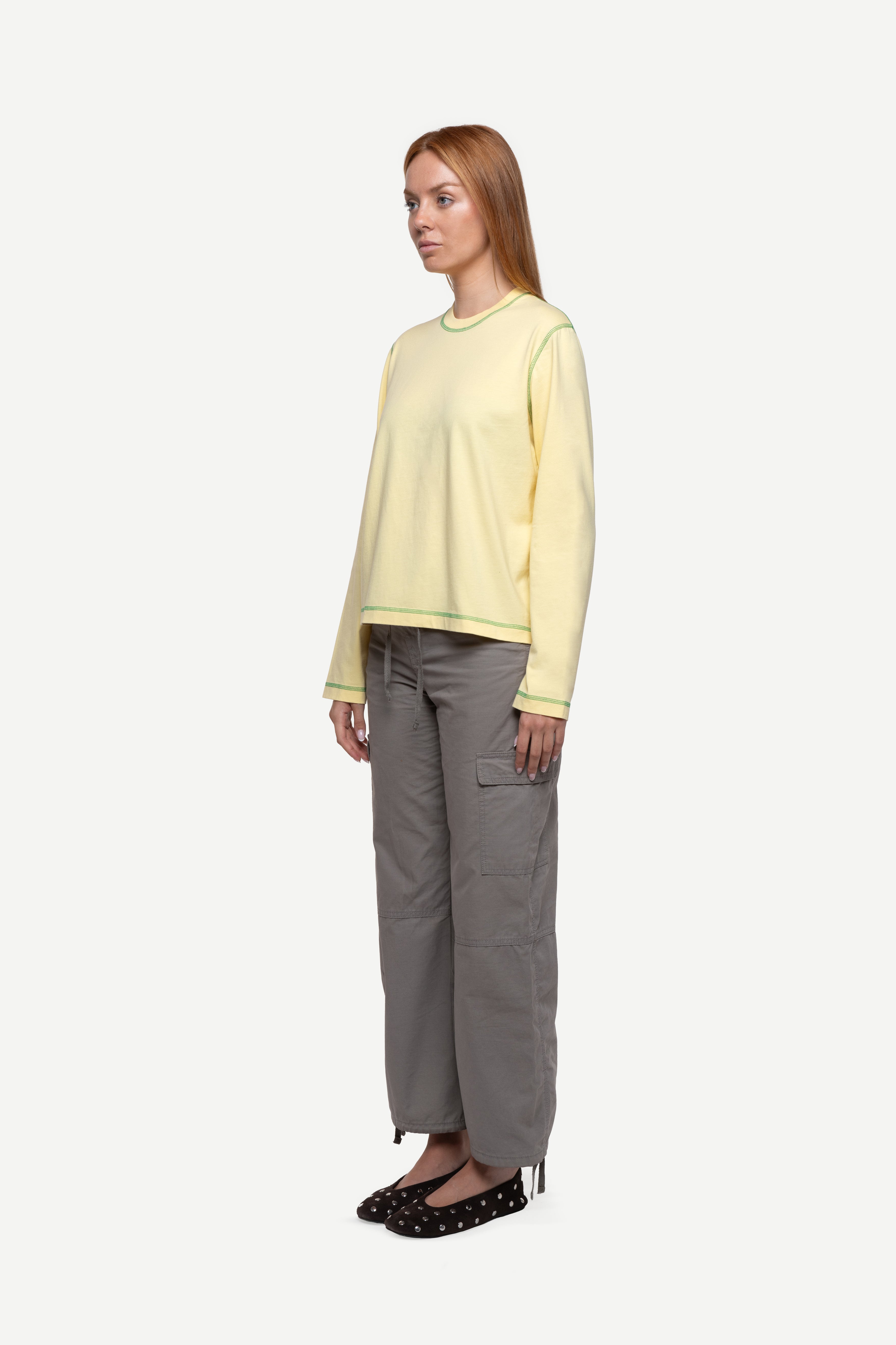 side view of woman wearing a regular fit pale yellow red long-sleeved t-shirt with contrast green over-lock stitching, cargo pants and ballet flats on a white background