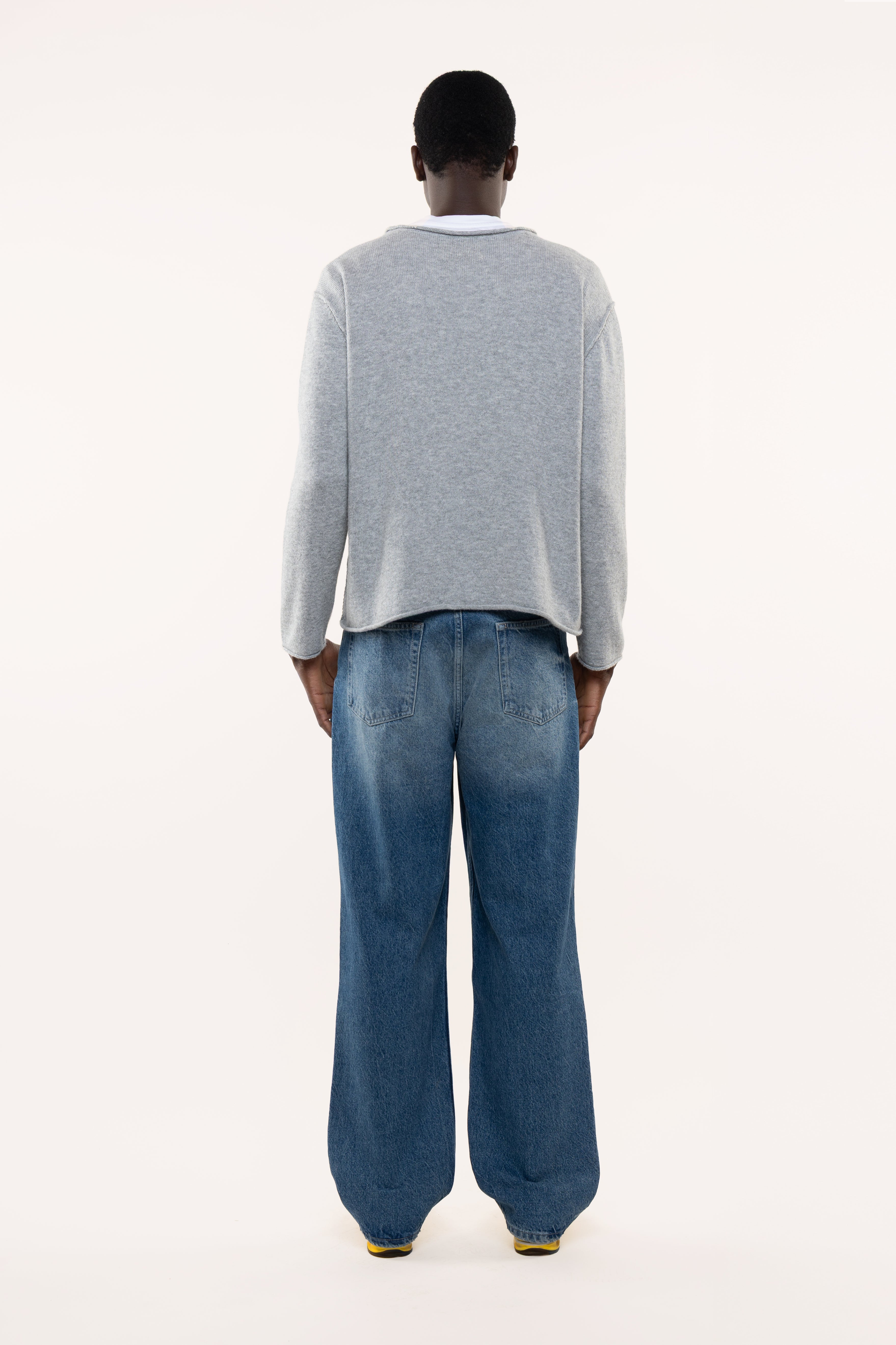 Back view of a man wearing a light gray sweater and blue jeans on a white background