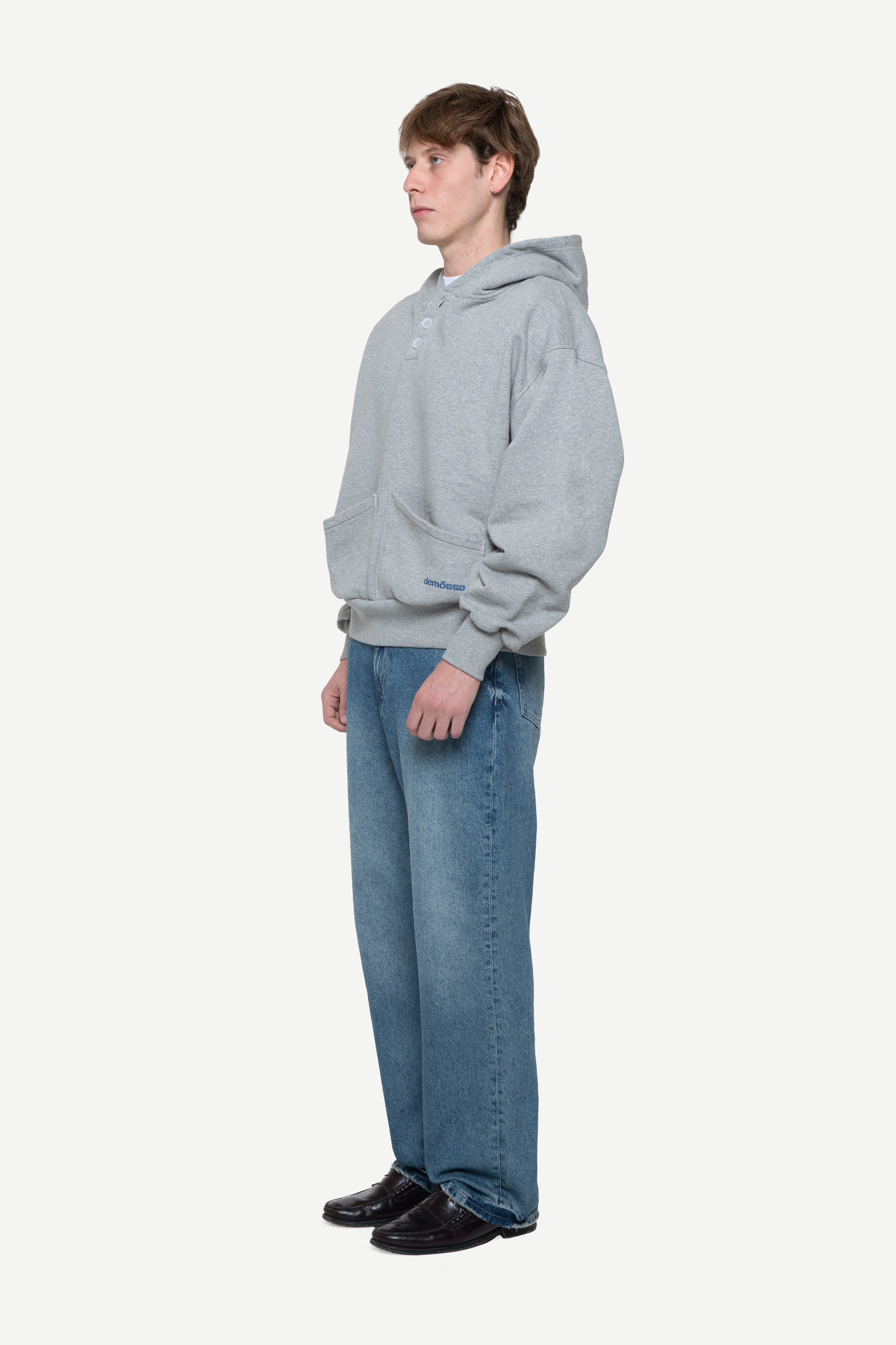 Side view of a man wearing a light grey hoodie with buttons on the neck and two front pockets and blue jeans on a white background