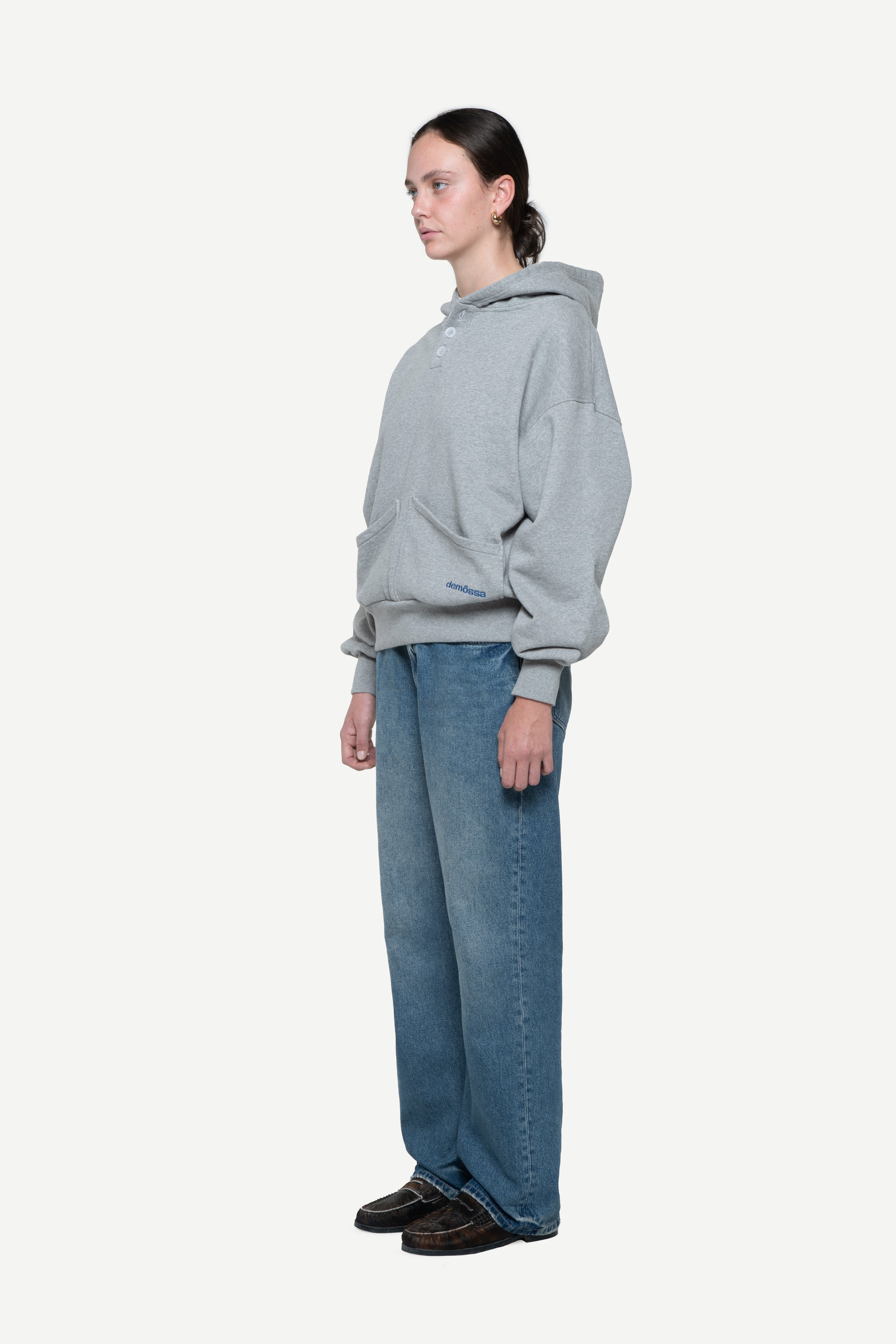 side view of a woman wearing a light grey hoodie with buttons on the neck and two front pockets and blue jeans on a white background