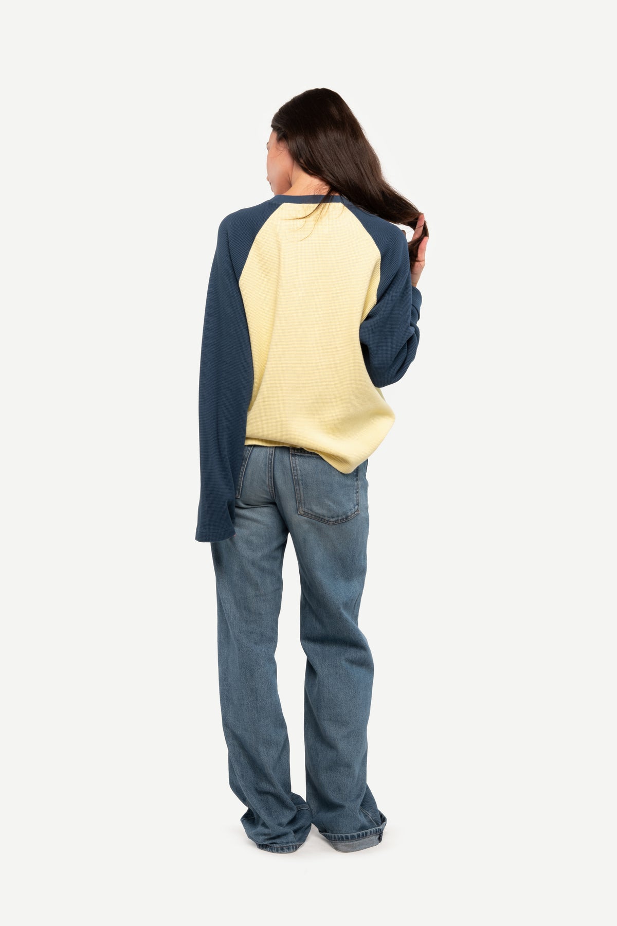 A model shown from the back wearing a two-tone raglan long-sleeve top with navy blue sleeves and shoulders and a pale yellow body, paired with blue jeans, against a plain light gray background.
