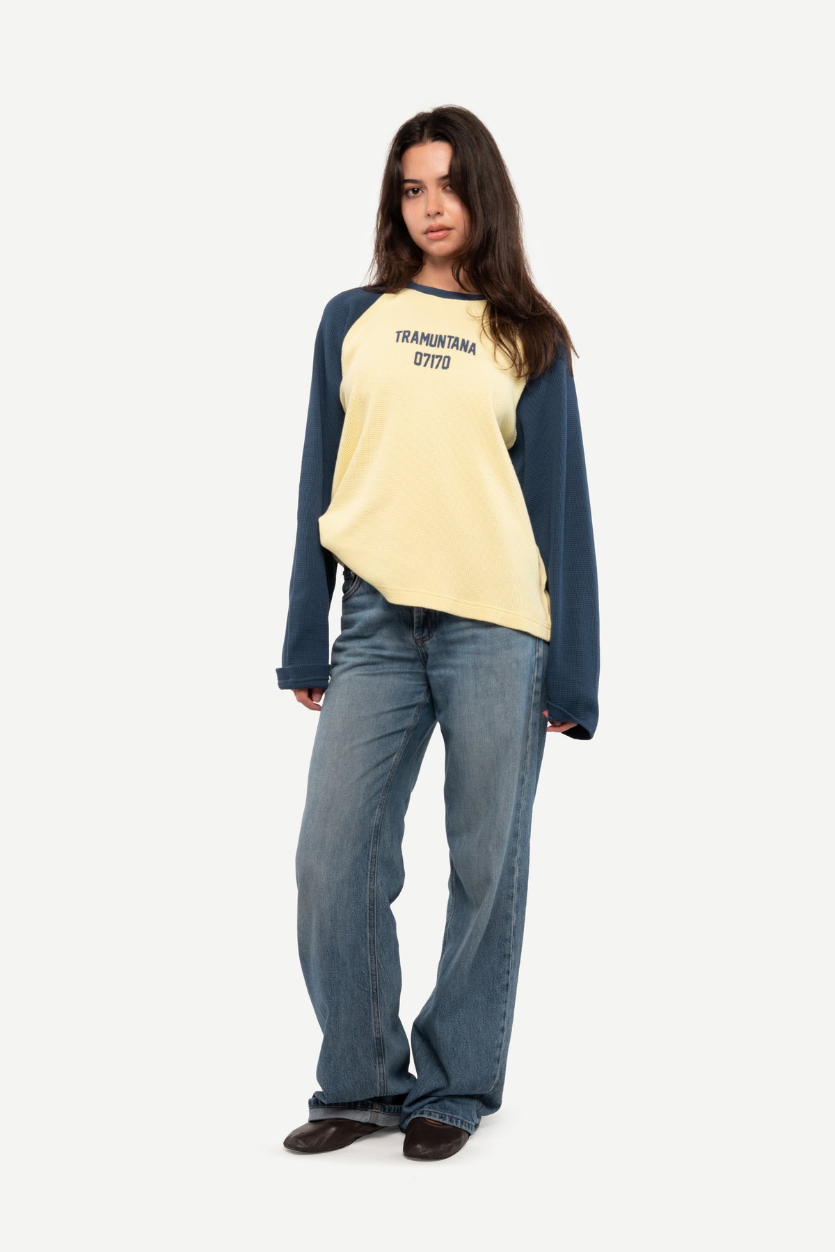 A woman modeling a long-sleeve raglan sweatshirt with a pale yellow body and dark navy sleeves, paired with wide-leg blue jeans and dark brown shoes, against a plain light gray background.
