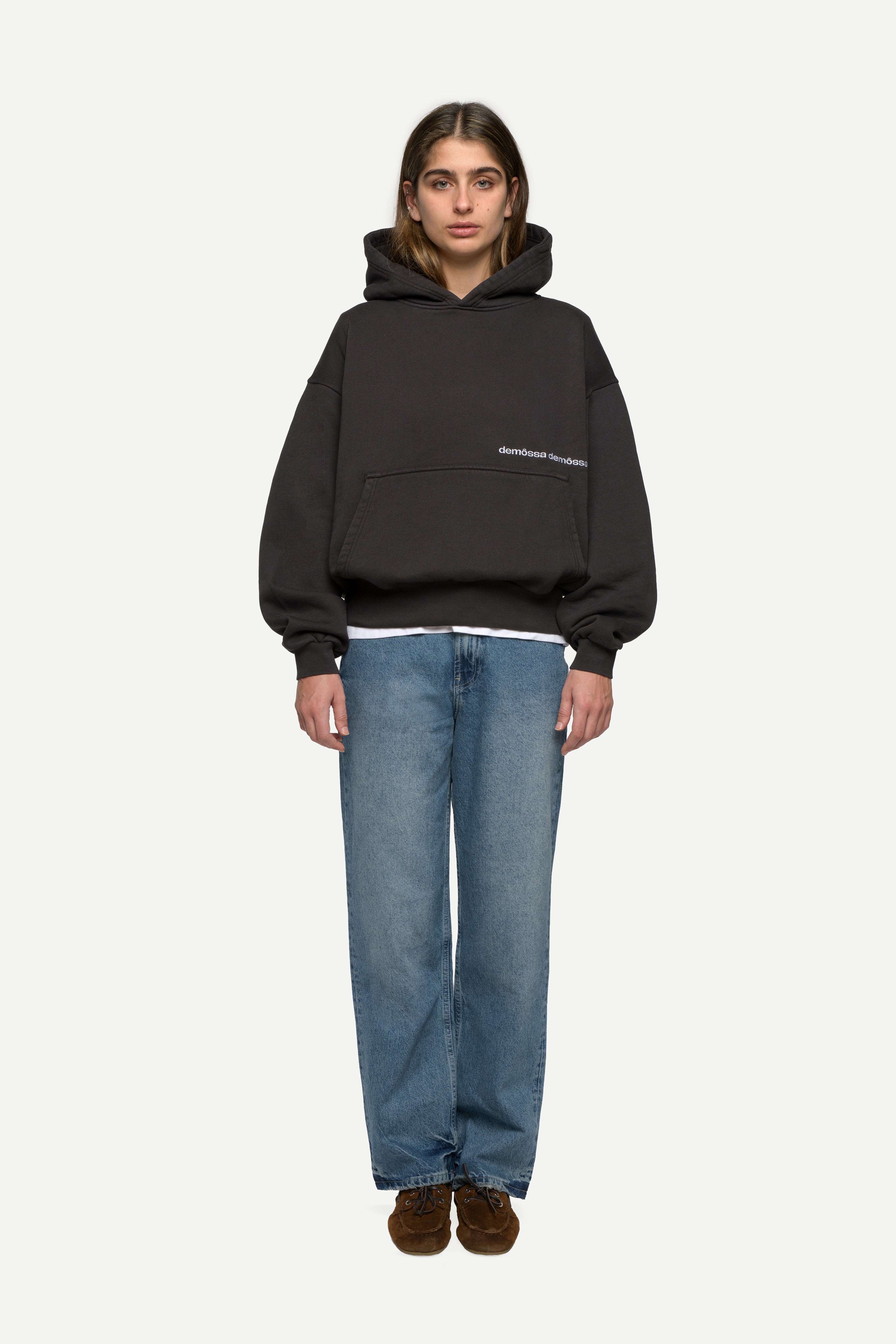 front view of woman wearing a black hoodie with double demôssa logo on the side and blue jeans on a white background