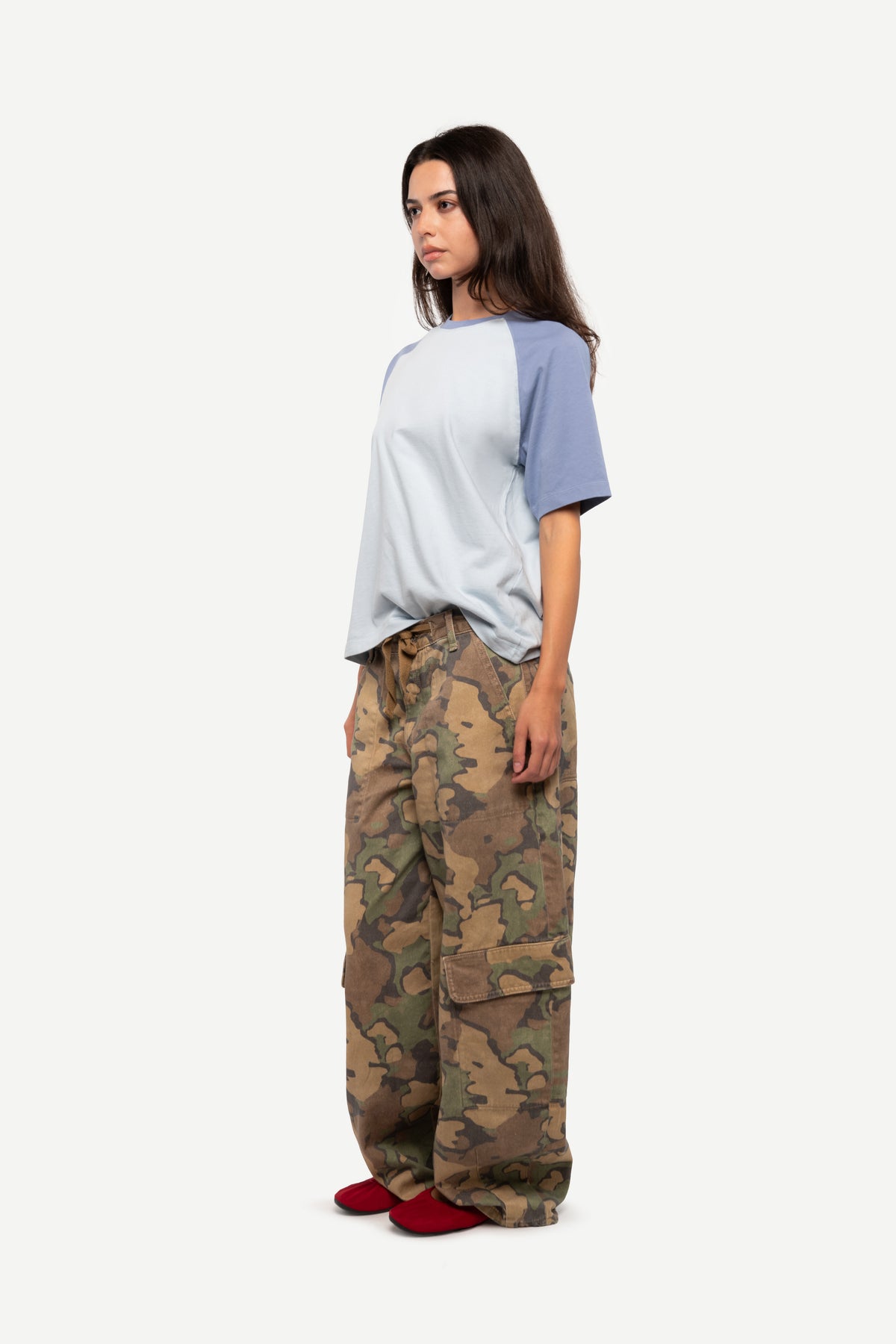 A woman stands in profile wearing a raglan-style short-sleeve t-shirt with a light gray body and muted blue sleeves, paired with loose-fit camouflage cargo pants and red slip-on shoes, against a plain light gray studio background.