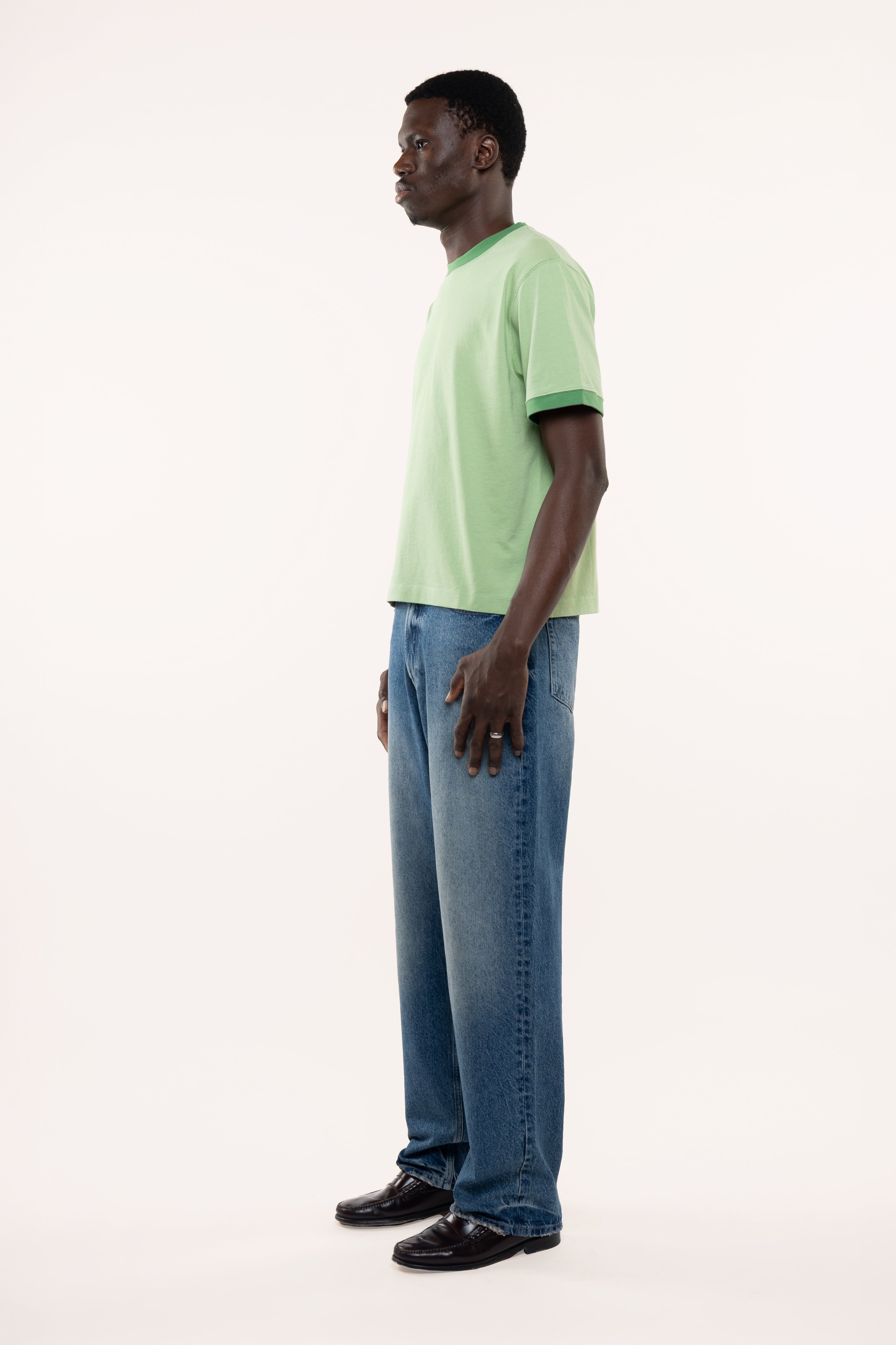 Side view of a man wearing a light green t-shirt and blue jeans standing against a white background
