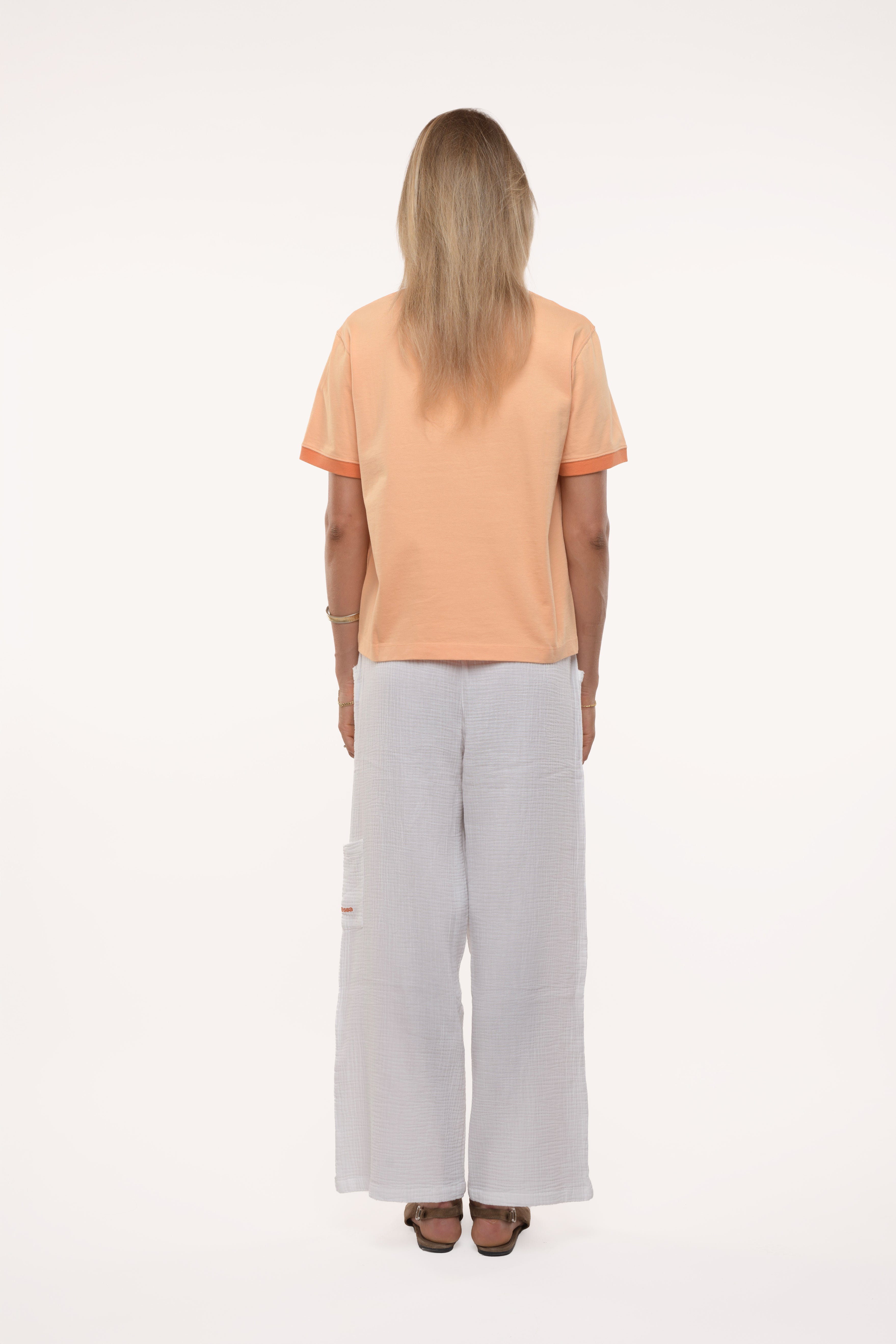 Back view of a woman wearing a peach-colored t-shirt and light gray pants on a white background