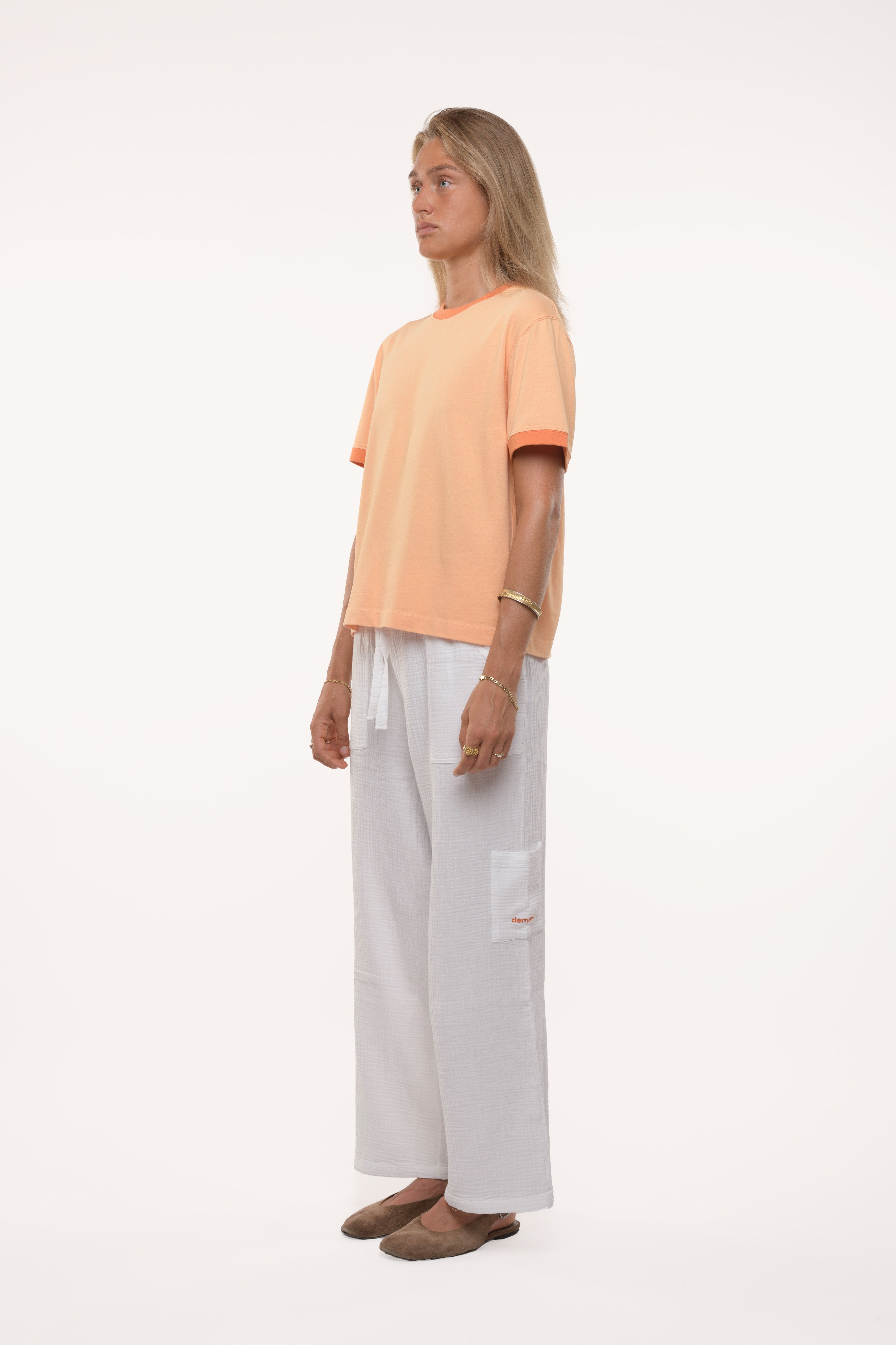 Side profile of a woman wearing a peach t-shirt and white pants on a white background