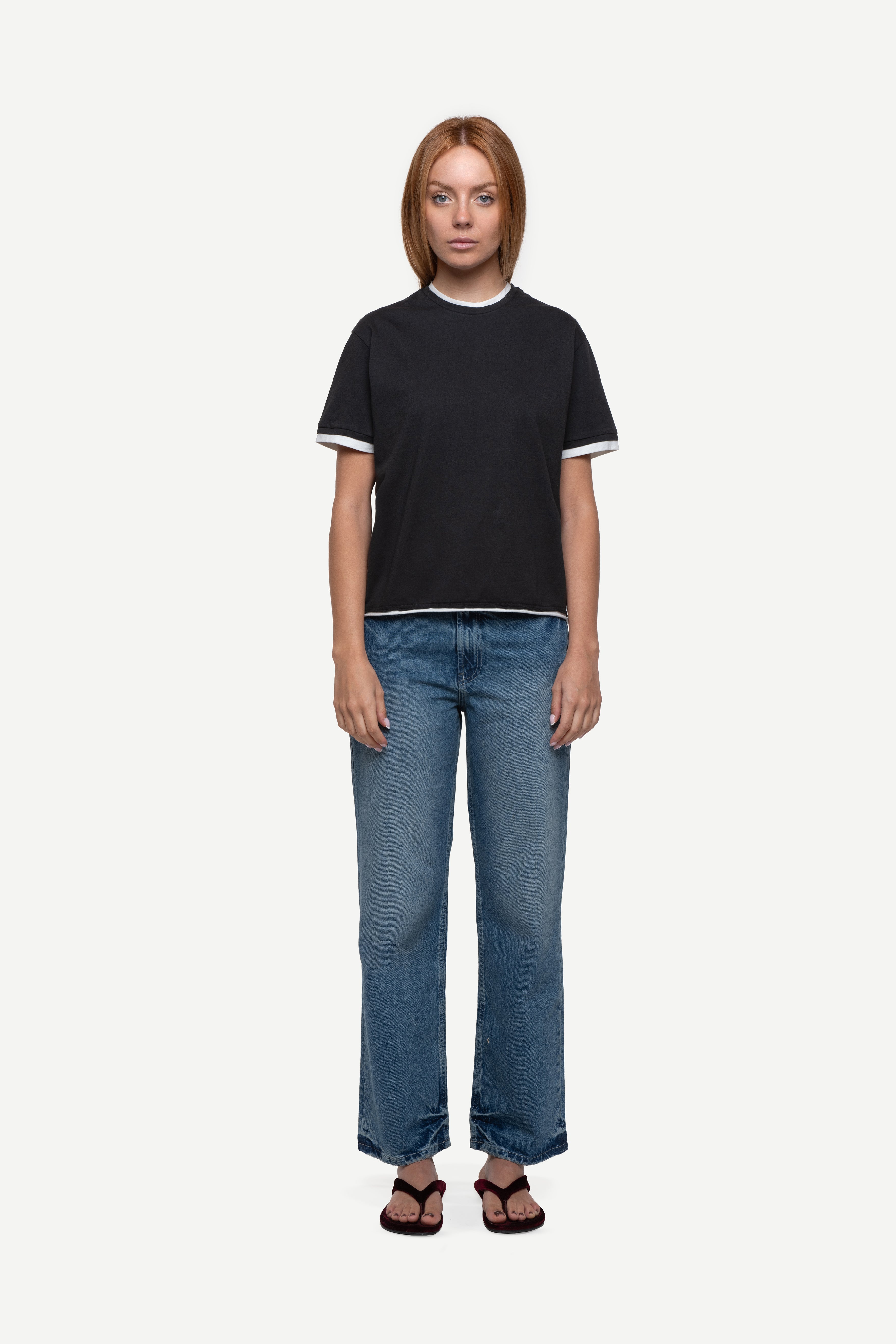 front view of woman wearing a regular fit black short-sleeved t-shirt with white double-layered neckline, sleeves and hem, blue jeans and flip flops on a white background
