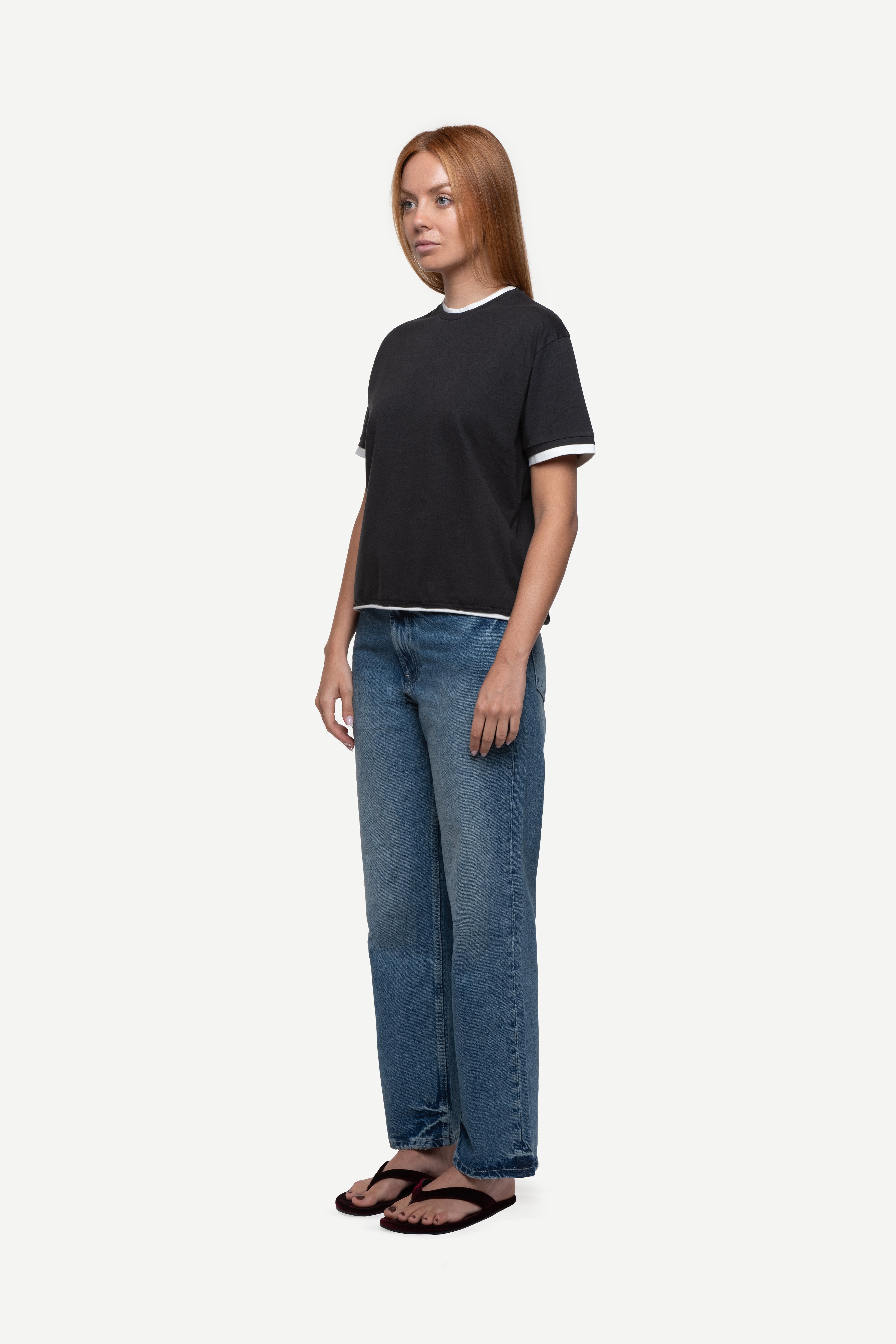 side view of woman wearing a regular fit black short-sleeved t-shirt with white double-layered neckline, sleeves and hem, blue jeans and flip flops on a white background