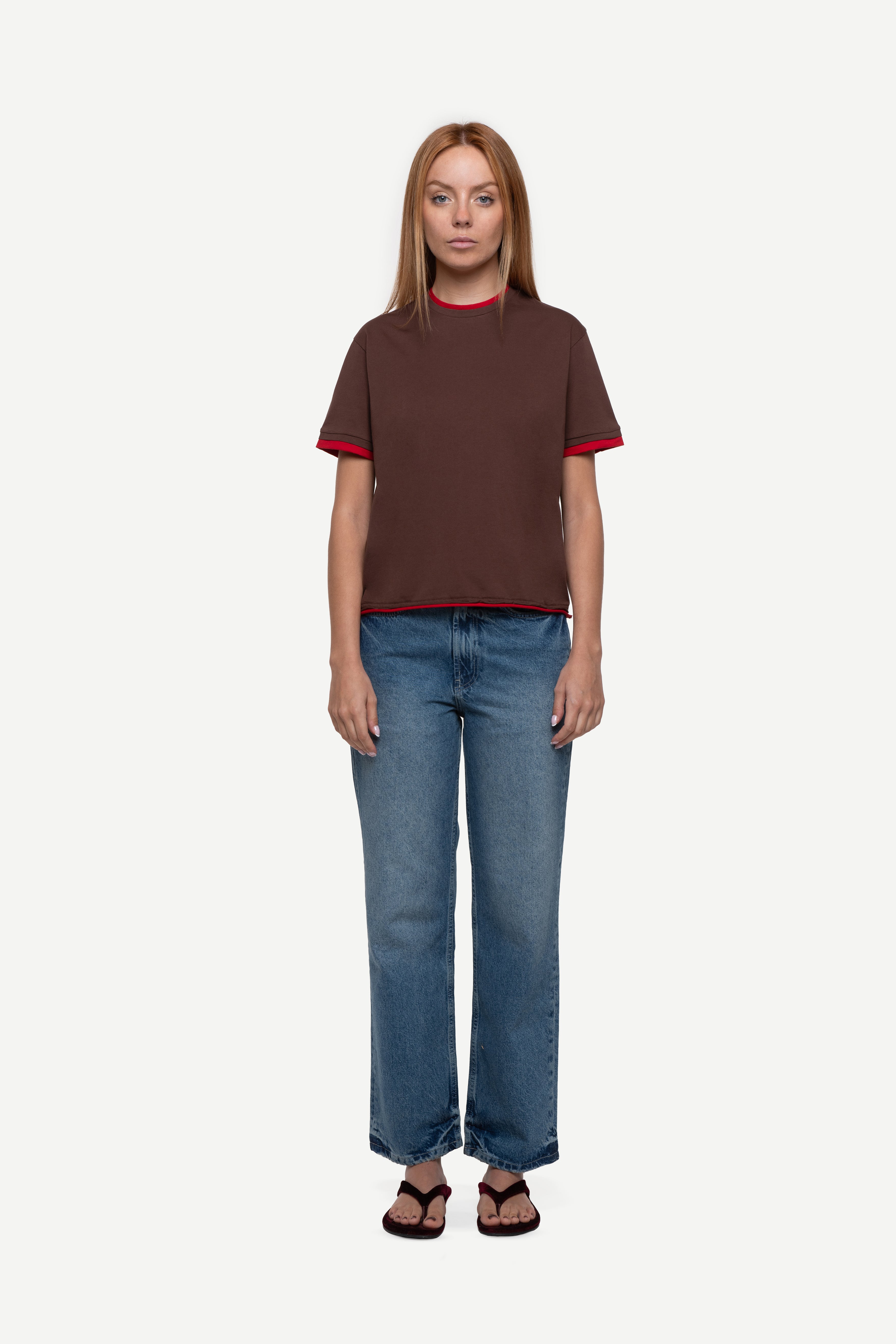 front view of woman wearing a regular fit brown short-sleeved t-shirt with red double-layered neckline, sleeves and hem, blue jeans and flip flops on a white background