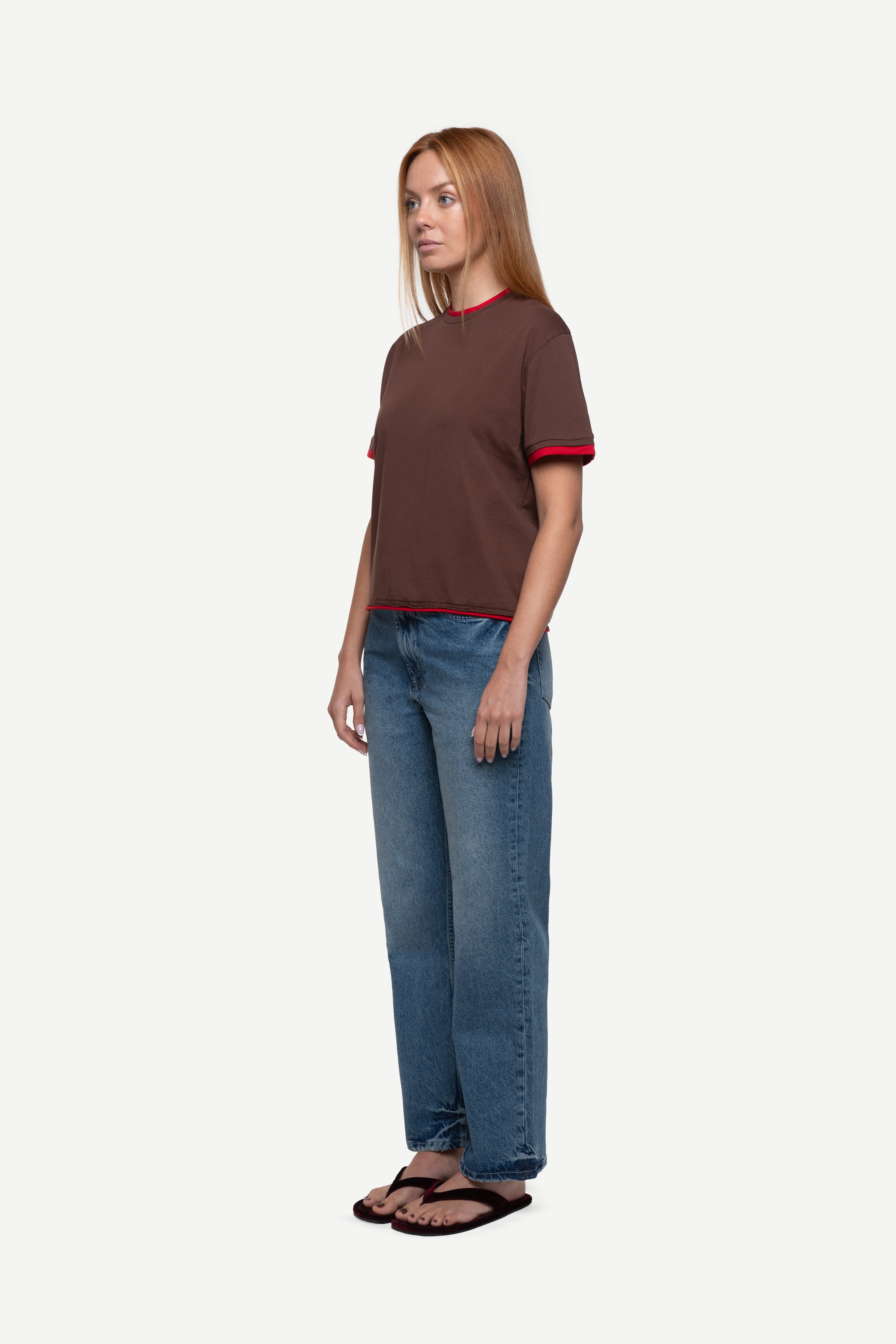 side view of woman wearing a regular fit brown short-sleeved t-shirt with red double-layered neckline, sleeves and hem, blue jeans and flip flops on a white background